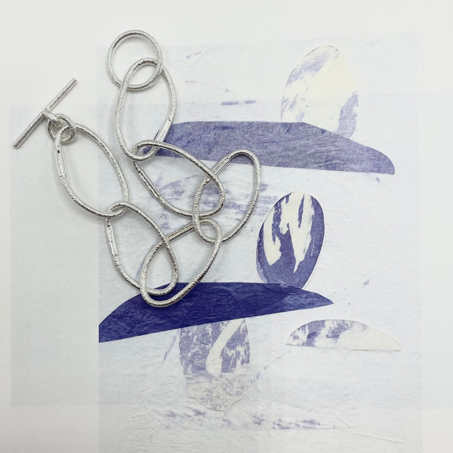 Versilbertes Armband aus Gliedern, die von Haargummis abgegossen sind, mit T-Verschluss, arrangiert auf einem grafischen Untergrund in Blau- und Weißtönen.