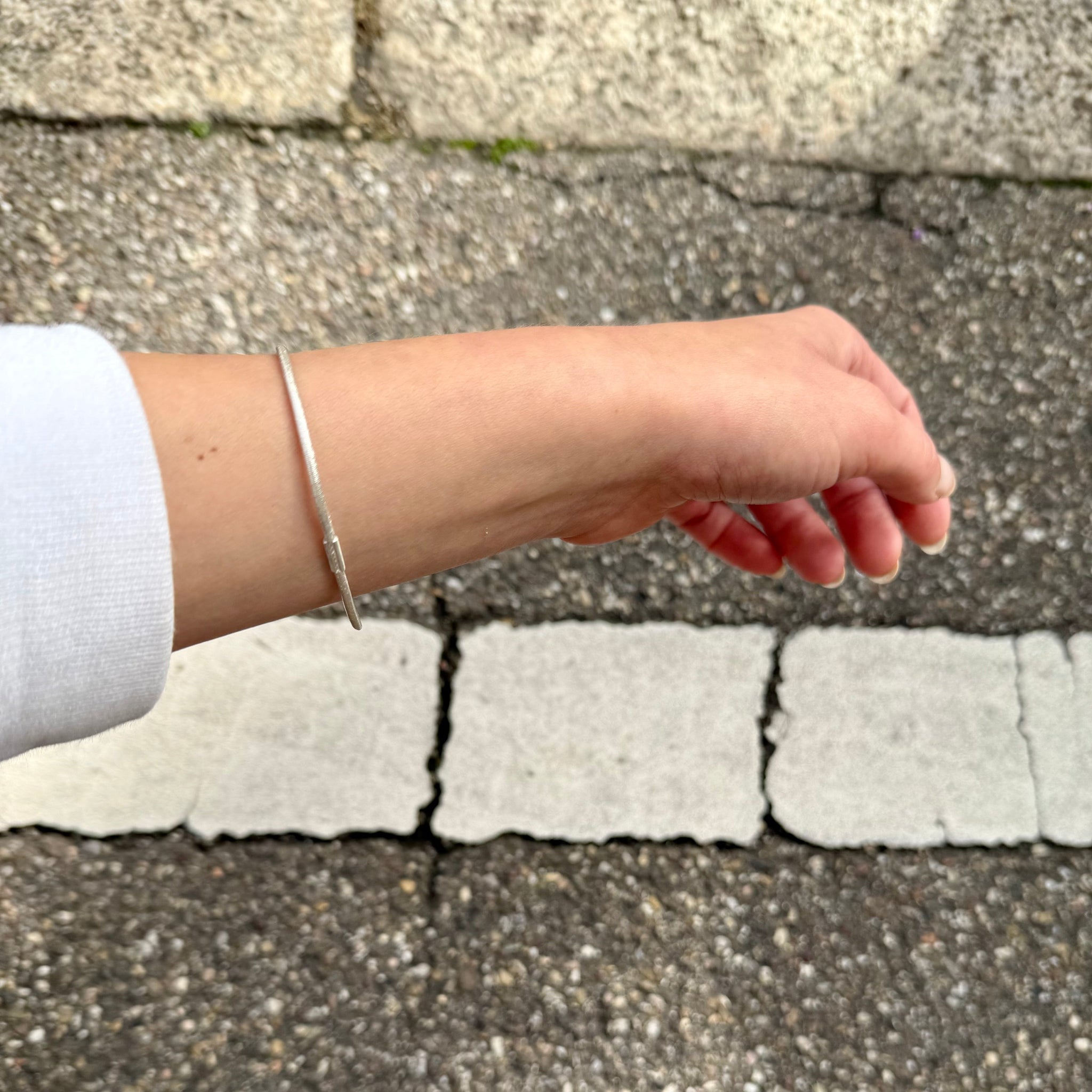 Silberner Armreif mit Haargummistruktur und Bördelung an der Schliesse, minimalistisch und handgefertigt, getragen an einem Handgelenk mit weissem Pulli.