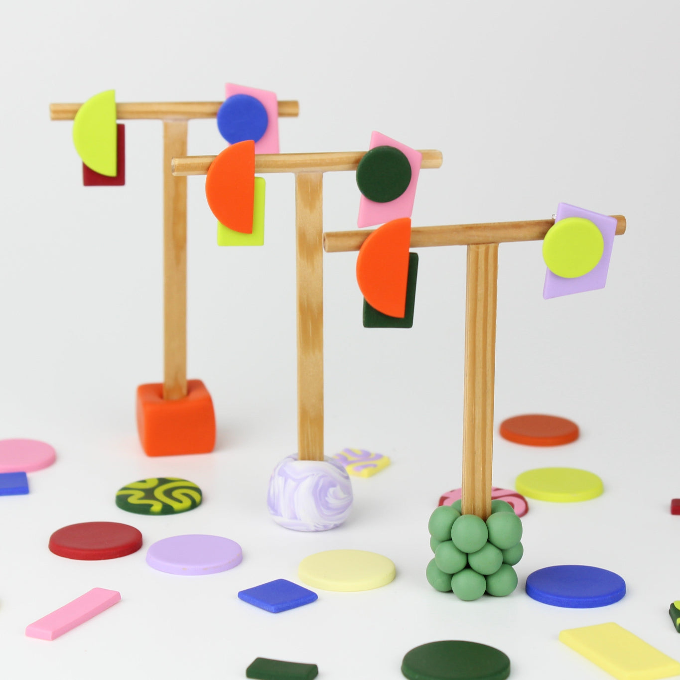 Geometrische Ohrhänger von Atelier Anda auf Schmuckständern arrangiert, umgeben von farbigen Polymerton-Elementen in verschiedenen Formen.