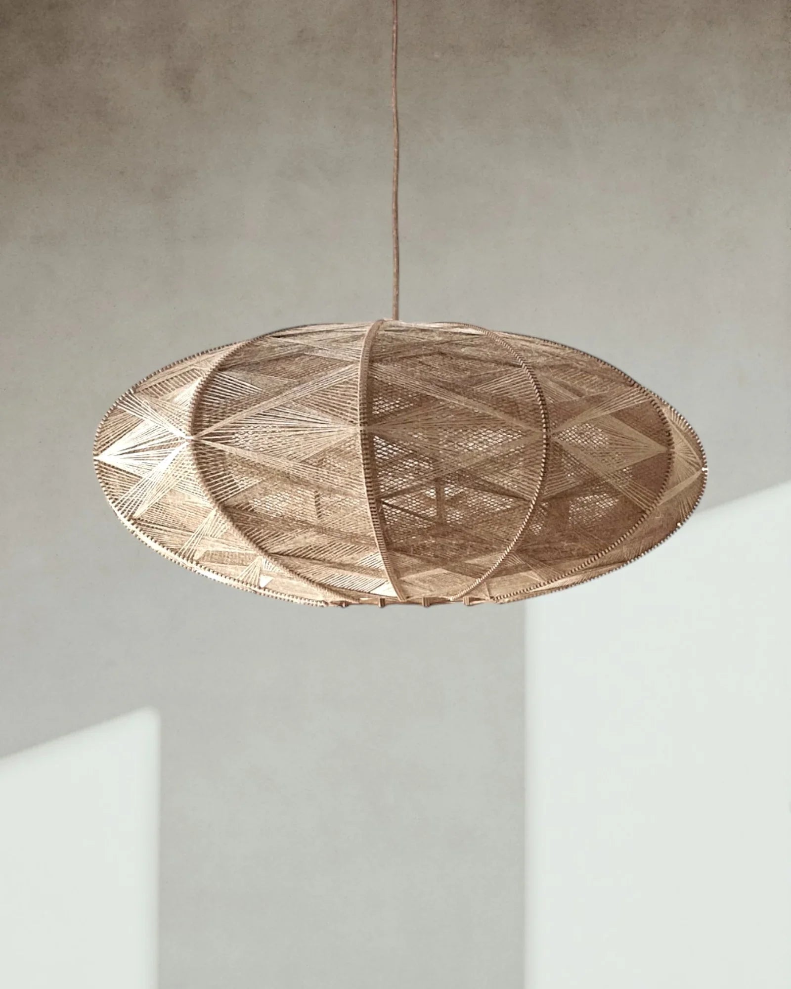eine elliptische Hängeleuchte aus Birkenholz umwebt mit beigem Leinengarn hängt vor einer sonnenbeschienenen Wand