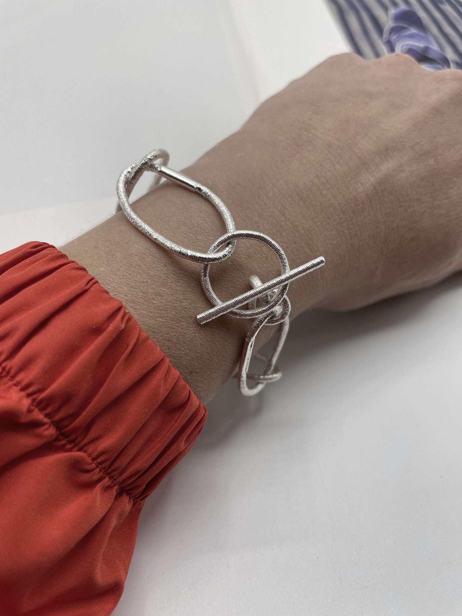 Versilbertes Gliederarmband mit Elementen, die Abgüsse von Haargummis sind, und T-Verschluss, getragen am Handgelenk einer Person mit orangefarbenem Ärmel.