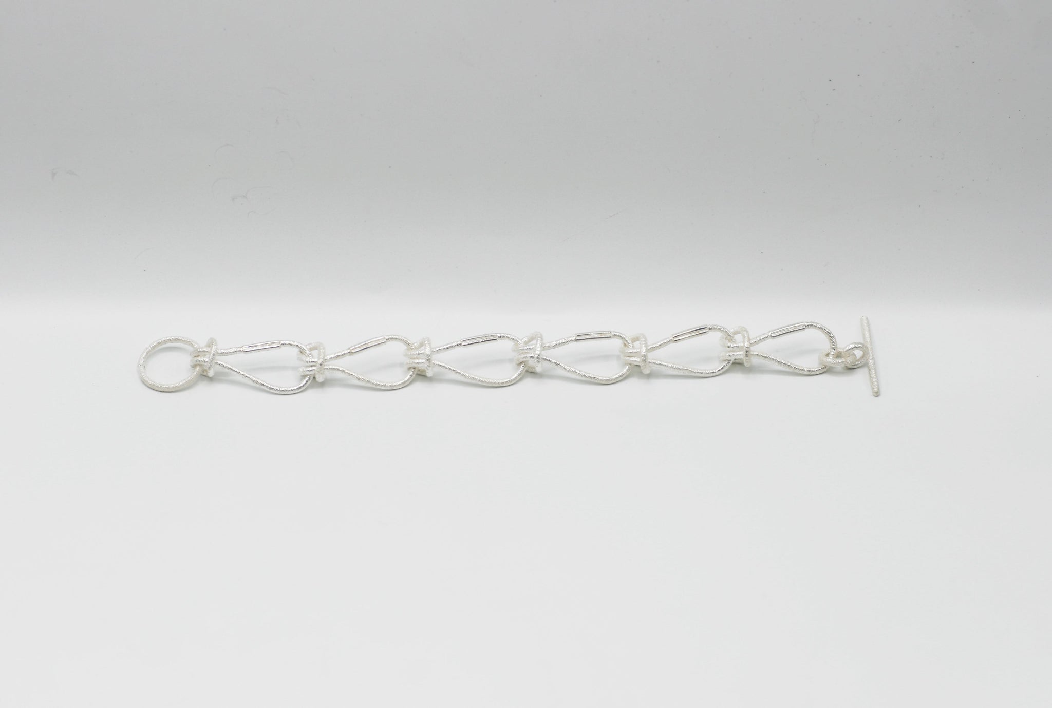 SilbernesArmband aus einzelnen, haargummi-ähnlichen, ineinander verknoteten Gliedern mit strukturierter Oberfläche und T-Verschluss, vor einem hellen Studiohintergrund