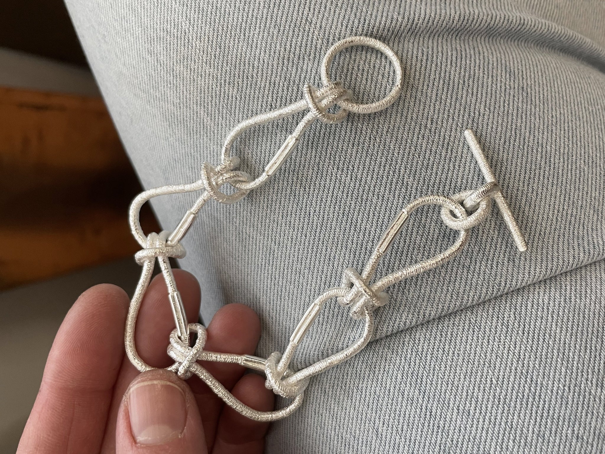 SilbernesArmband aus einzelnen, haargummi-ähnlichen, ineinander verknoteten Gliedern mit strukturierter Oberfläche und T-Verschluss, locker in einer Hand gehalten vor hellem Jeansstoff.