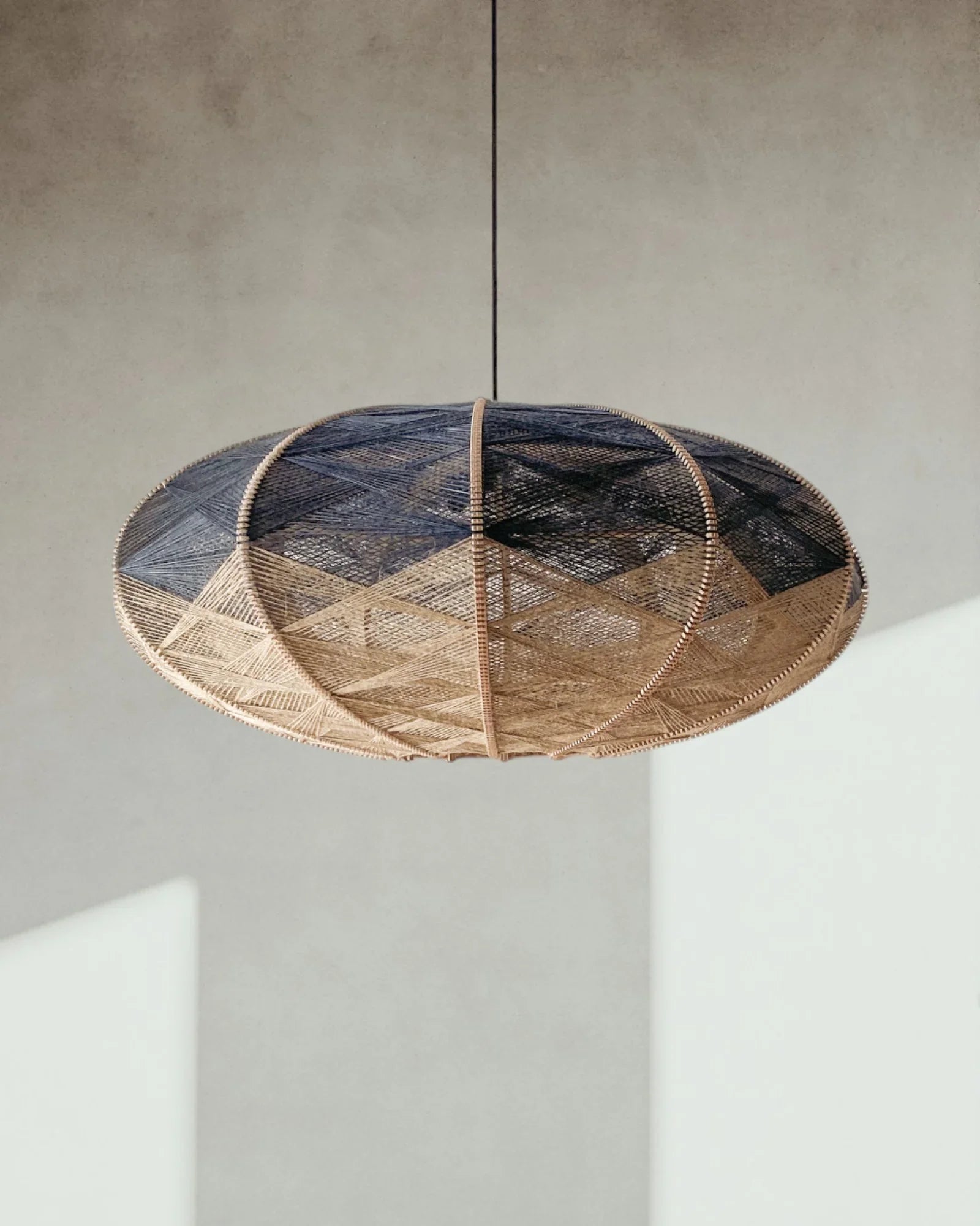 eine elliptische Hängeleuchte aus Birkenholz umwebt mit Leinengarn in Beige und Anthrazit hängt vor einer sonnenbeschienenen Wand