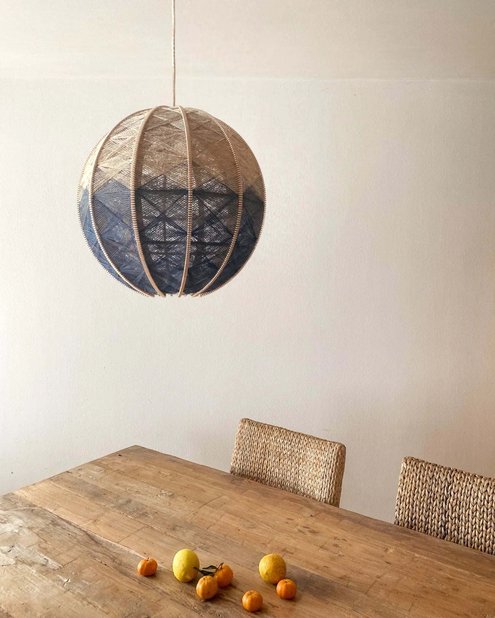 eine runde Hängeleuchte aus Birkenholz umwebt mit Leinengarn in Beige und Anthrazit hängt über einem Holztisch mit Zitrusfrüchten