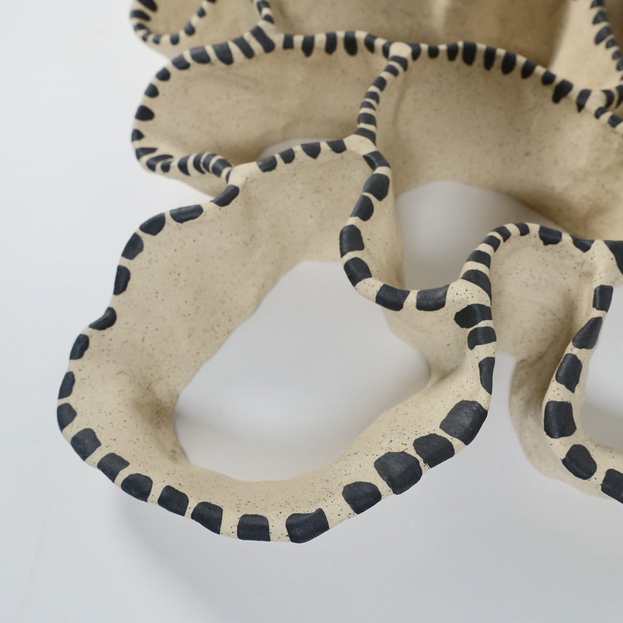 Detailansicht einer keramischen Wandskulptur mit offenem, durchbrochenem Aufbau, angelehnt an eine Korallenform; sandfarbene Oberfläche mit unregelmäßig geschwungenen Formen, entlang der Kanten mit rhythmisch gesetzten, schwarzen Pinselstrichen akzentuiert.