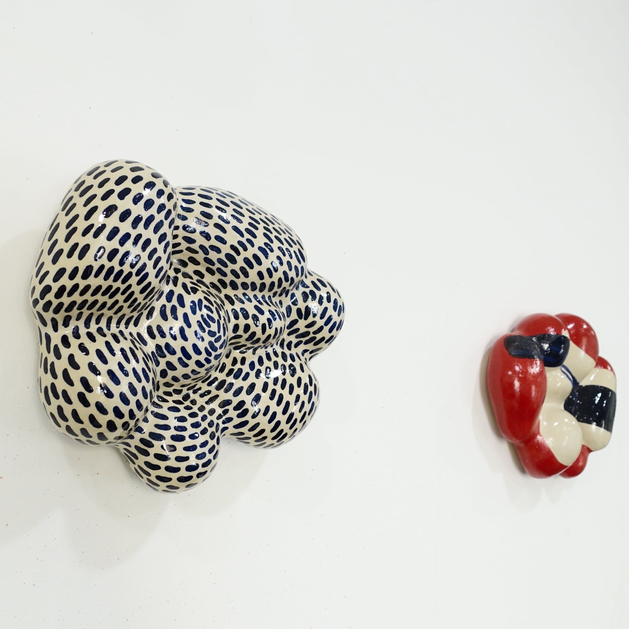 Zwei Wandskulpture aus Keramik mit organisch gewölbter, wolkenähnlicher Form; eine mit hellem Grundmaterial, dicht überzogen mit gleichmäßig verteilten dunkelblauen Pinselstrichen; die andere mit hellem Grundtin, roten Flächen udn gestischen dunkellauen und schwarzen, abstrakten Formen.