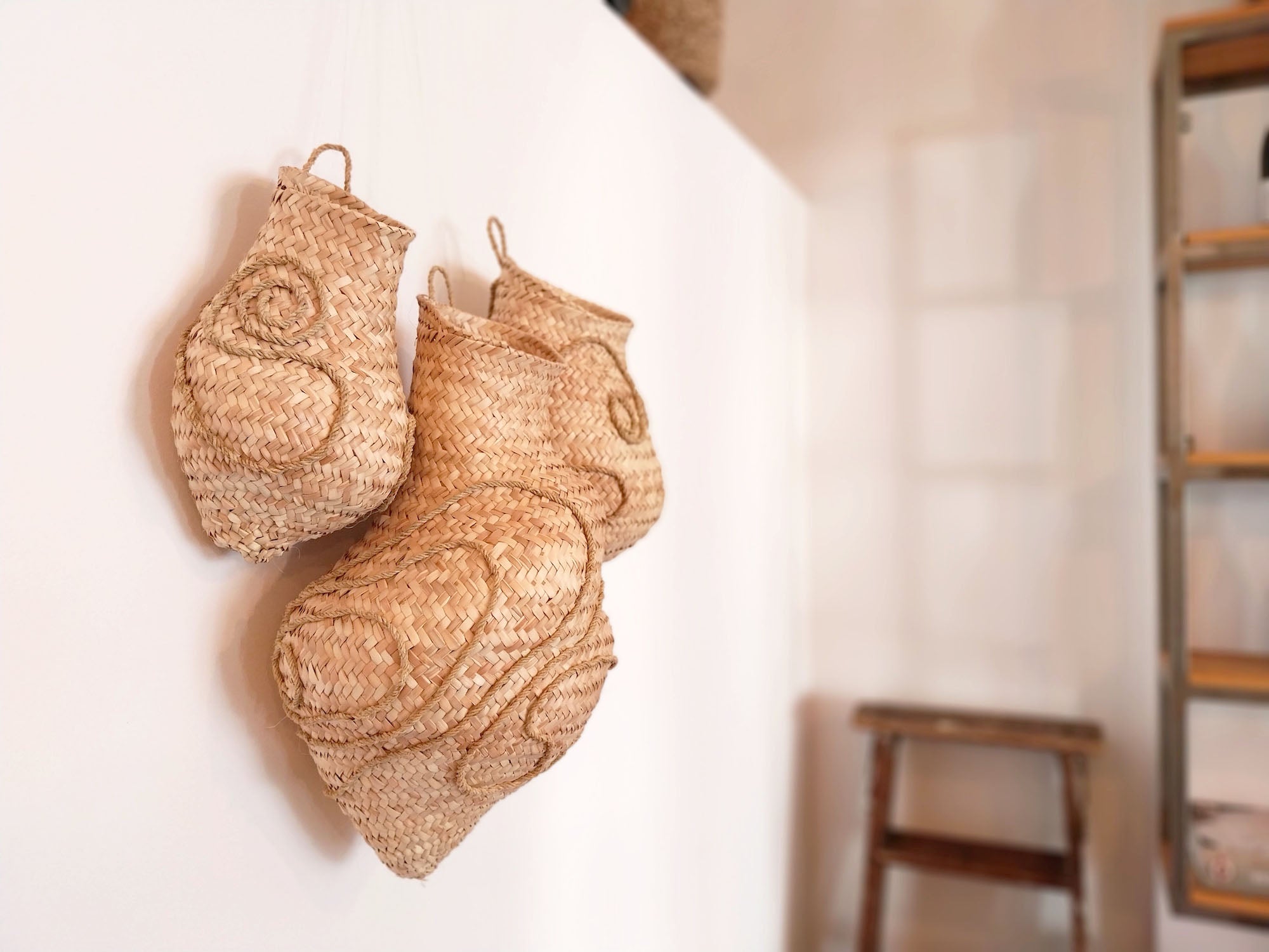 Wandskulptur aus drei handgeflochtenen Körben aus hellen Palmwedeln, organisch geformt und mit reliefartigen Spiral- und Linienmustern versehen, dicht nebeneinander an einer weißen Wand montiert; die natürliche Faserstruktur und warmen Beigetöne stehen im Kontrast zum ruhigen, minimalistischen Raum.