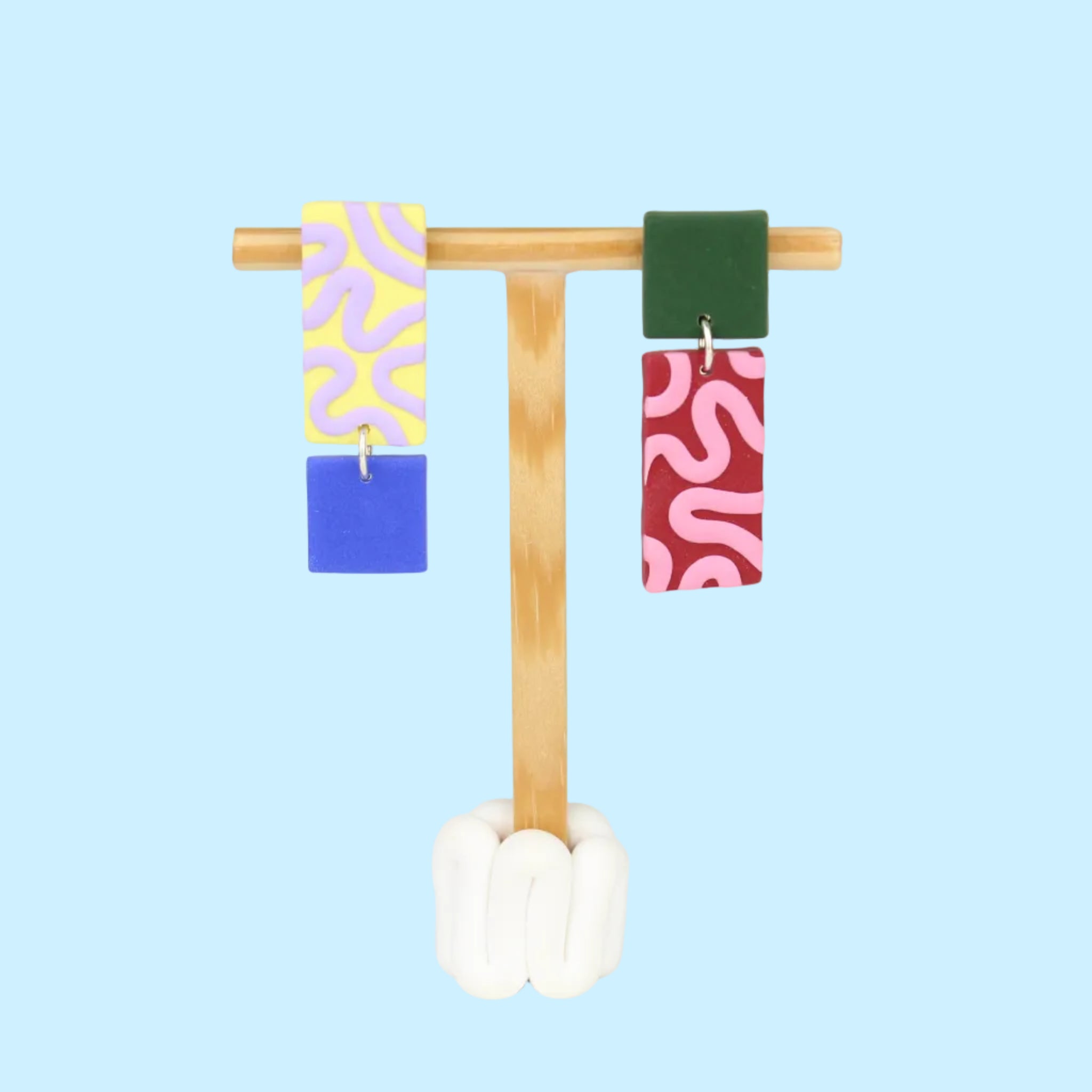 Asymmetrische Ohrringe aus Polymerton mit rechteckigen Elementen und grafischen Mustern in Gelb, Rot, Flieder, Blau und Grün, präsentiert auf einem hölzernen Schmuckständer.