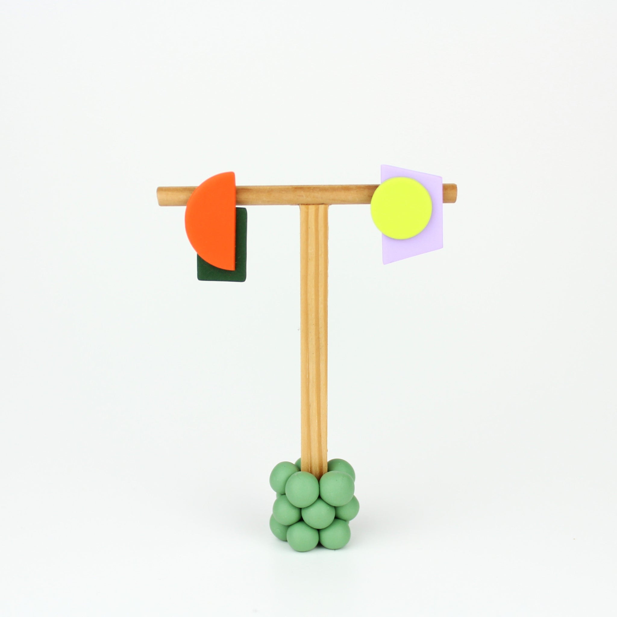 Asymmetrische Ohrringe aus Polymerton mit grafischen Formen in Orange, Dunkelgrün, Neongelb und Flieder, präsentiert auf einem schlichten Schmuckständer vor pastellfarbenem Hintergrund.