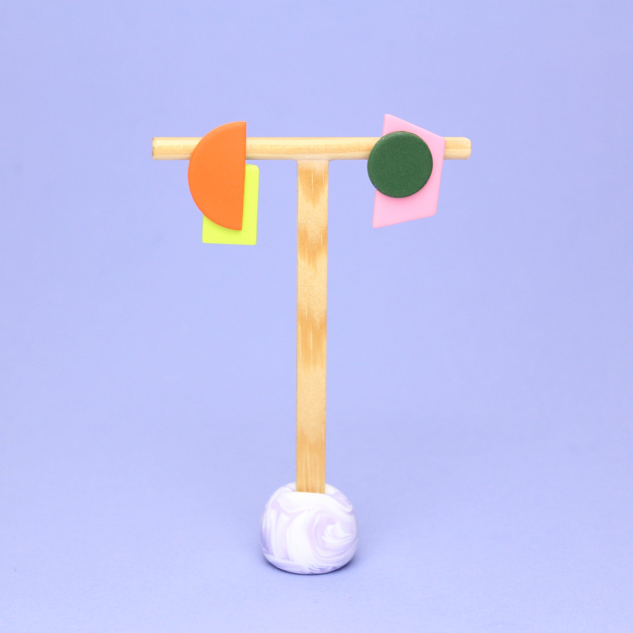 Asymmetrische Ohrringe aus Polymerton mit grafischen Formen in Orange, Grün, Gelb und Rosa, präsentiert auf einem schlichten Schmuckständer vor pastellfarbenem Hintergrund.