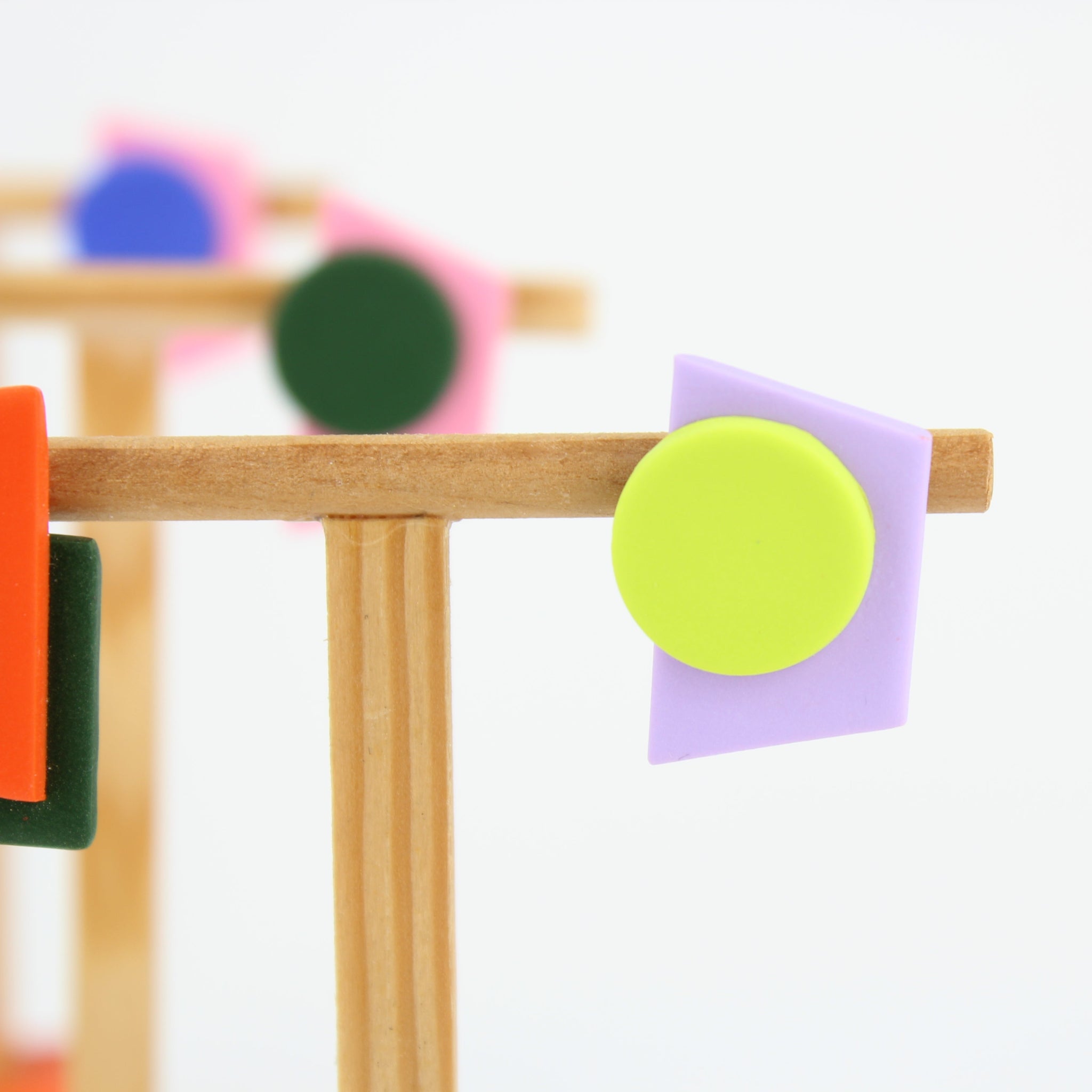 Asymmetrische Ohrringe aus Polymerton mit grafischen Formen in Orange, Dunkelgrün, Neongelb und Flieder, präsentiert auf einem schlichten Schmuckständer vor pastellfarbenem Hintergrund.