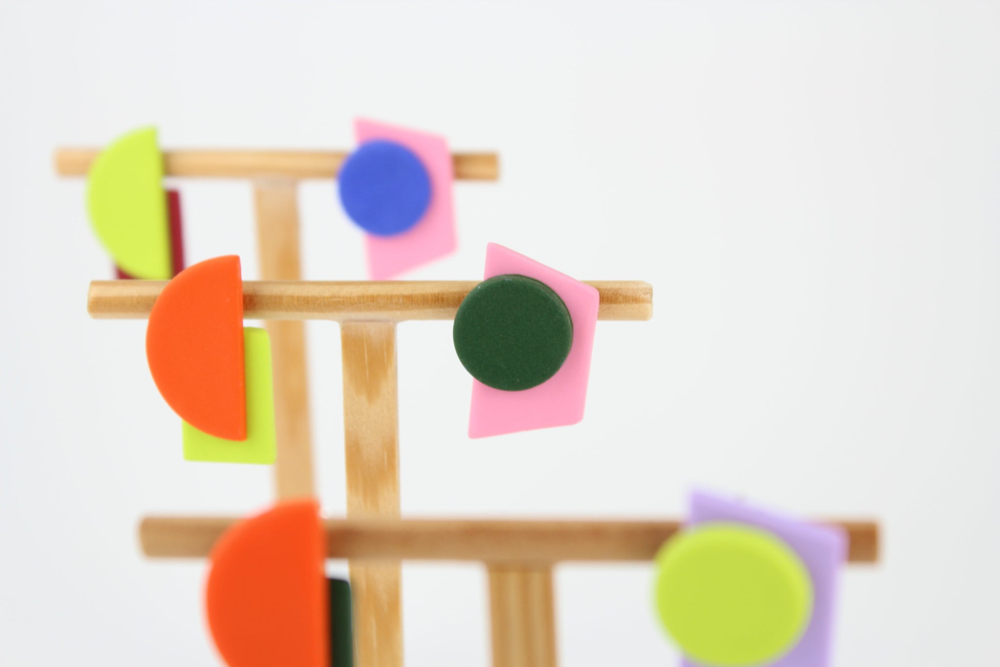 Mehrere asymmetrische Ohrringe aus Polymerton mit geometrischen Formen in kräftigen Farben, arrangiert auf hölzernen Schmuckständern vor hellem Hintergrund.