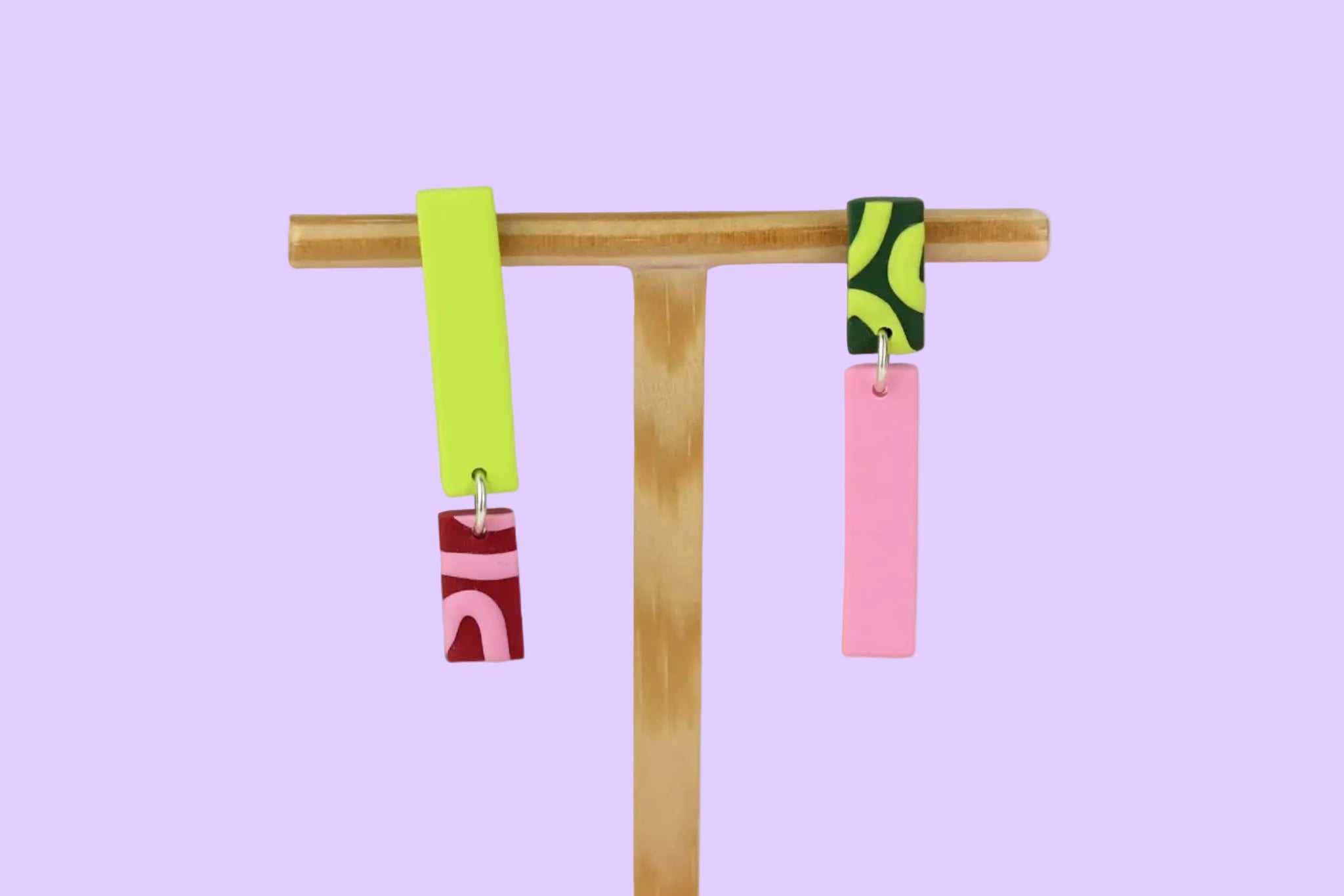 Asymmetrische Ohrringe aus Polymerton mit schmalen rechteckigen Elementen und grafischen Mustern in Rot, Rosa, Grün und Neongelb, präsentiert auf einem hölzernen Schmuckständer.