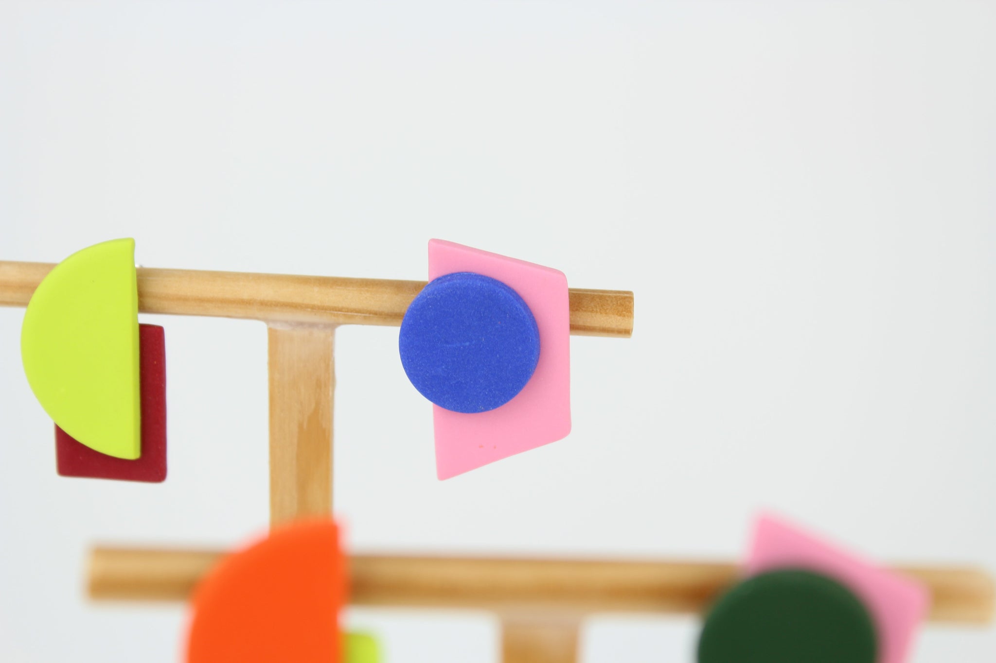 Mehrere asymmetrische Ohrringe aus Polymerton mit geometrischen Formen in kräftigen Farben, arrangiert auf hölzernen Schmuckständern vor hellem Hintergrund.