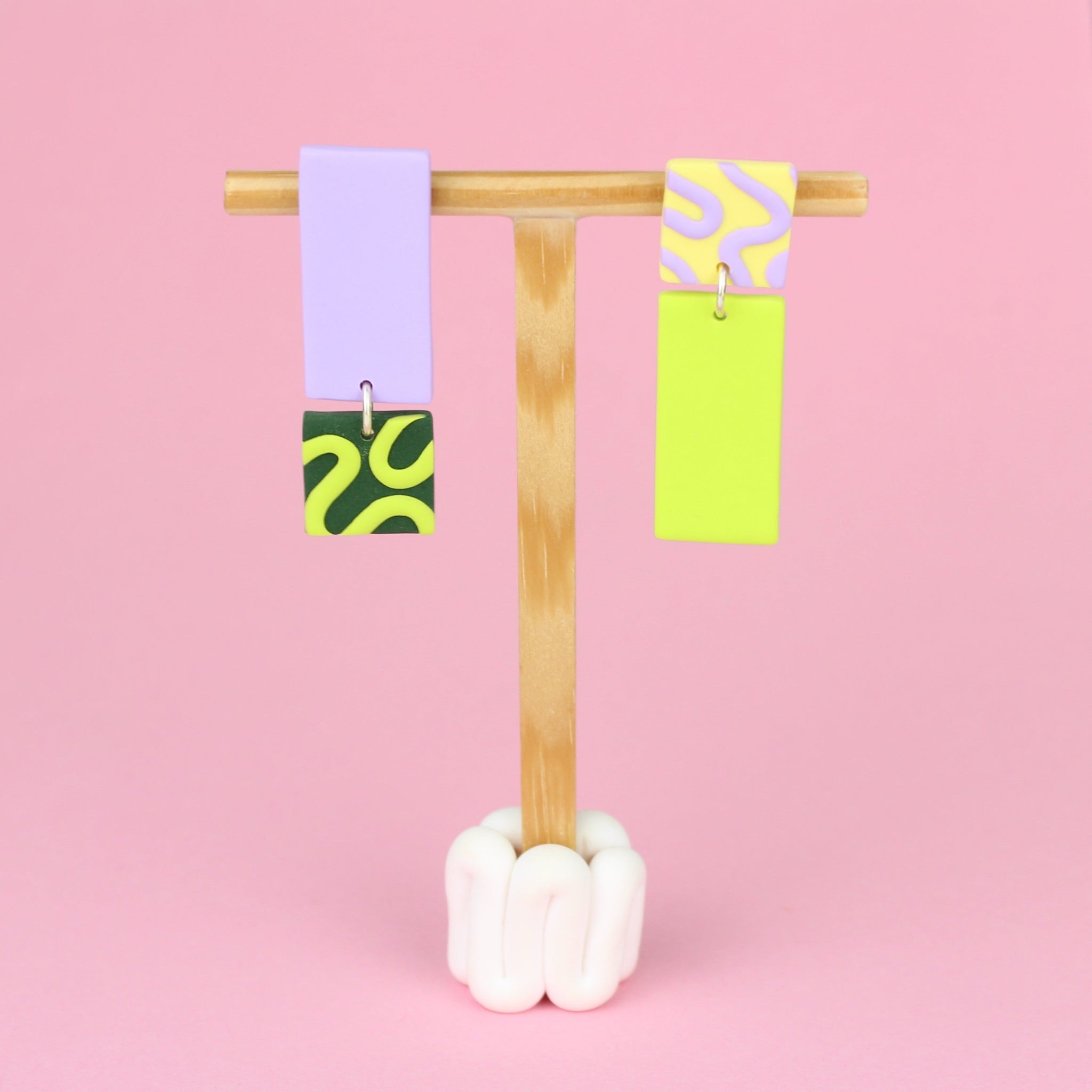 Asymmetrische Ohrringe aus Polymerton mit rechteckigen Elementen und grafischen Mustern in Flied, präsentiert auf einem hölzernen Schmuckständer.