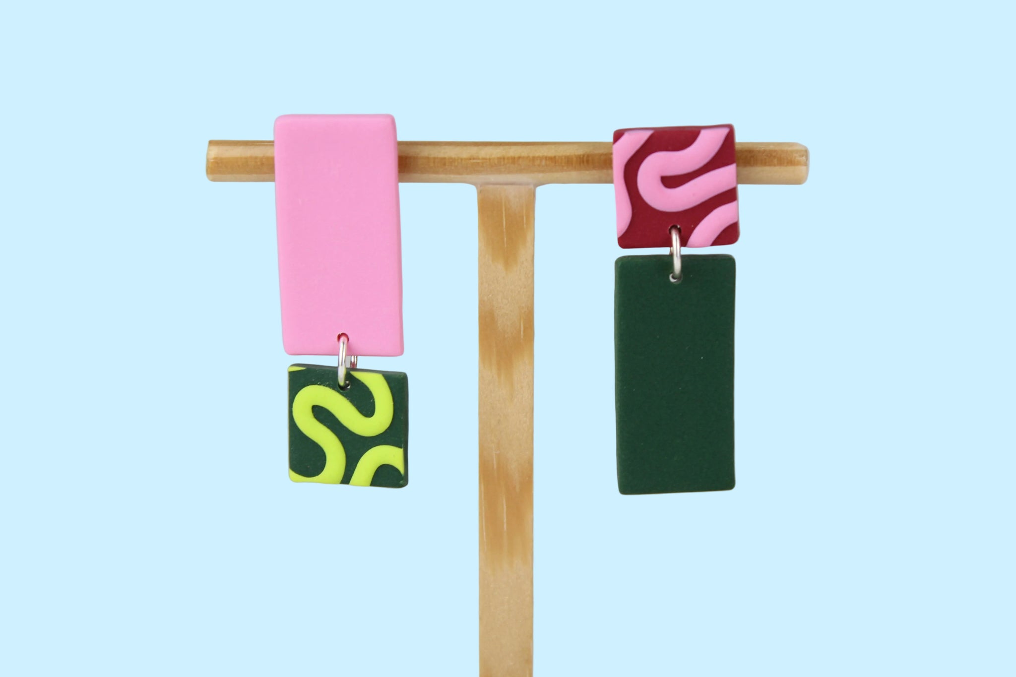 Asymmetrische Ohrringe aus Polymerton mit rechteckigen Elementen und grafischen Mustern in Rosa und Grün, präsentiert auf einem hölzernen Schmuckständer vor hellblauem Hintergrund.