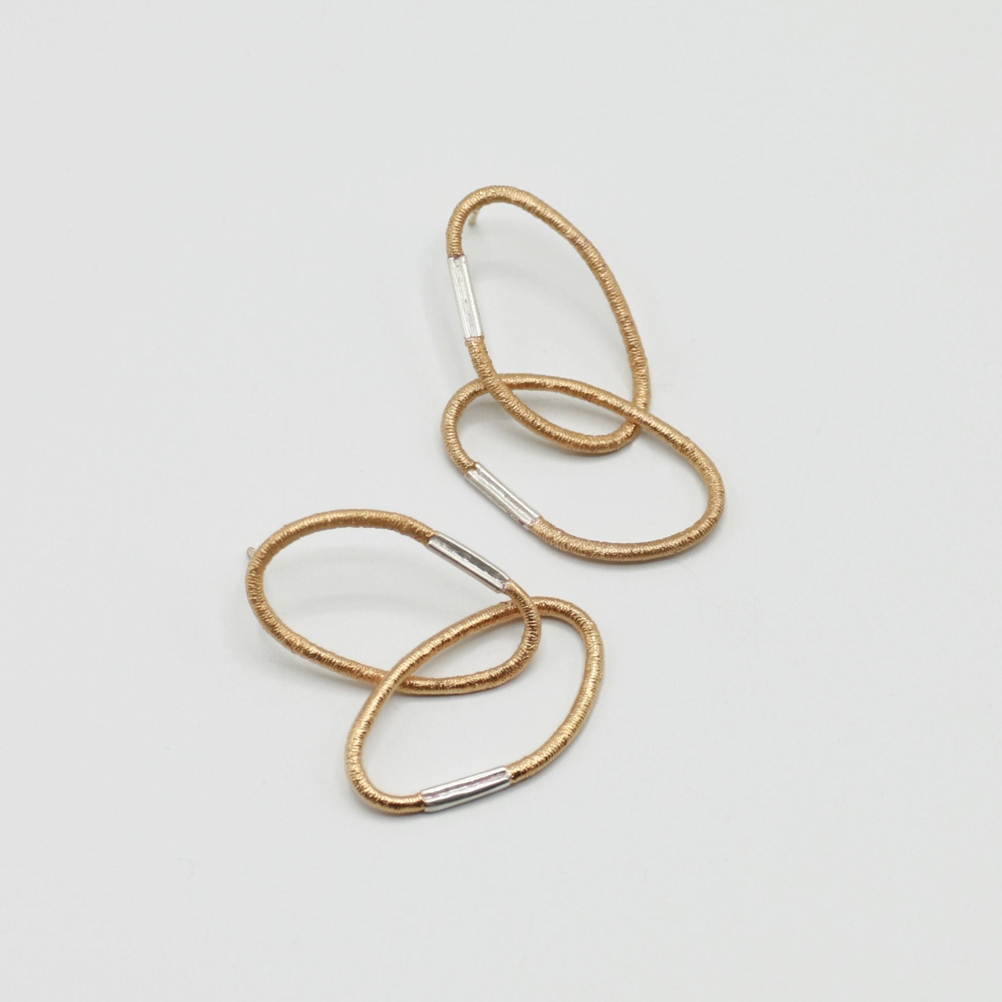 Ein Paar Ohrringe aus je zwei ovalen Gliedern mit charakteristischer Haargummistruktur und Bördelungsdetail an der Schliesse, minimalistisch und handgefertigt. Vergoldetes Silber mit silberner Schliesse, arrangiert auf hellem Hintergrund.
