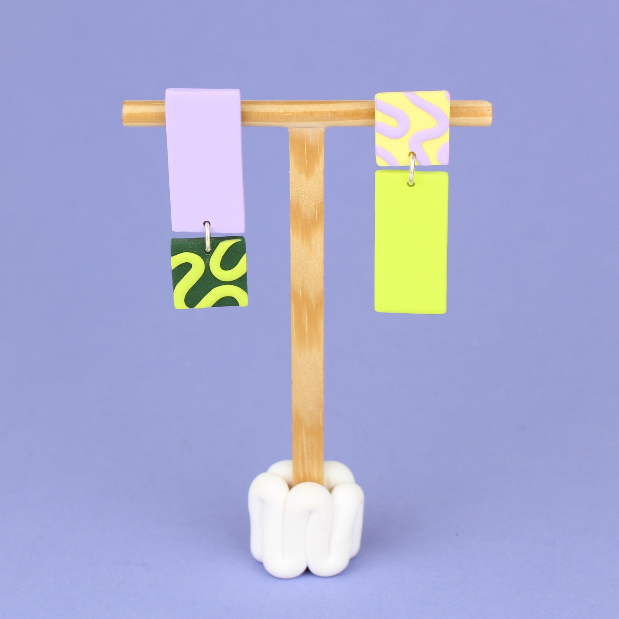 Asymmetrische Ohrringe aus Polymerton mit rechteckigen Elementen und grafischen Mustern in Flied, präsentiert auf einem hölzernen Schmuckständer.