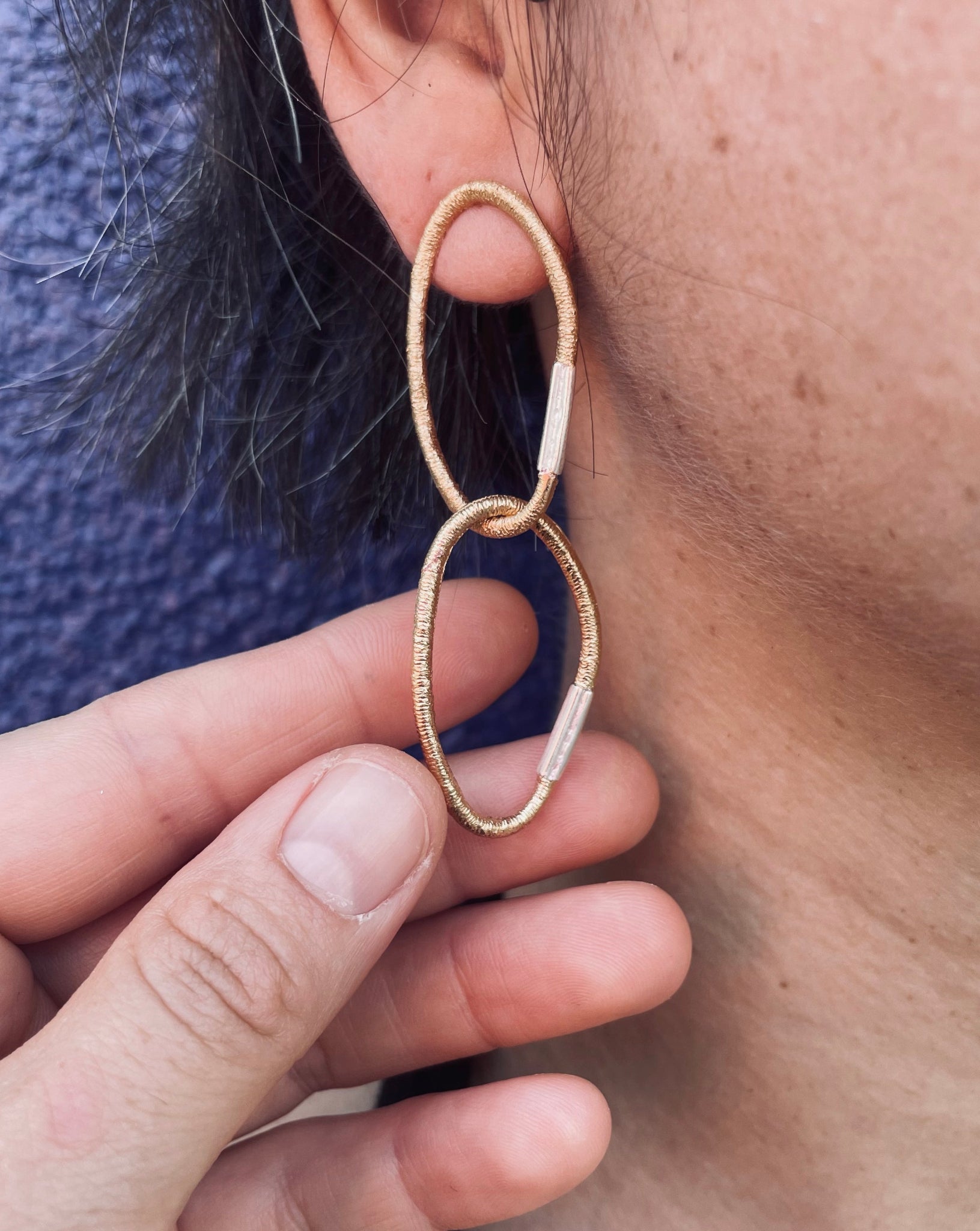 Vergoldete Silber-Ohrringe aus zwei ovalen Gliedern mit charakteristischer Haargummistruktur und silbernem Bördelungsdetail an der Schliesse, minimalistisch und handgefertigt, am Ohr getragen vor blauem Hintergrund.