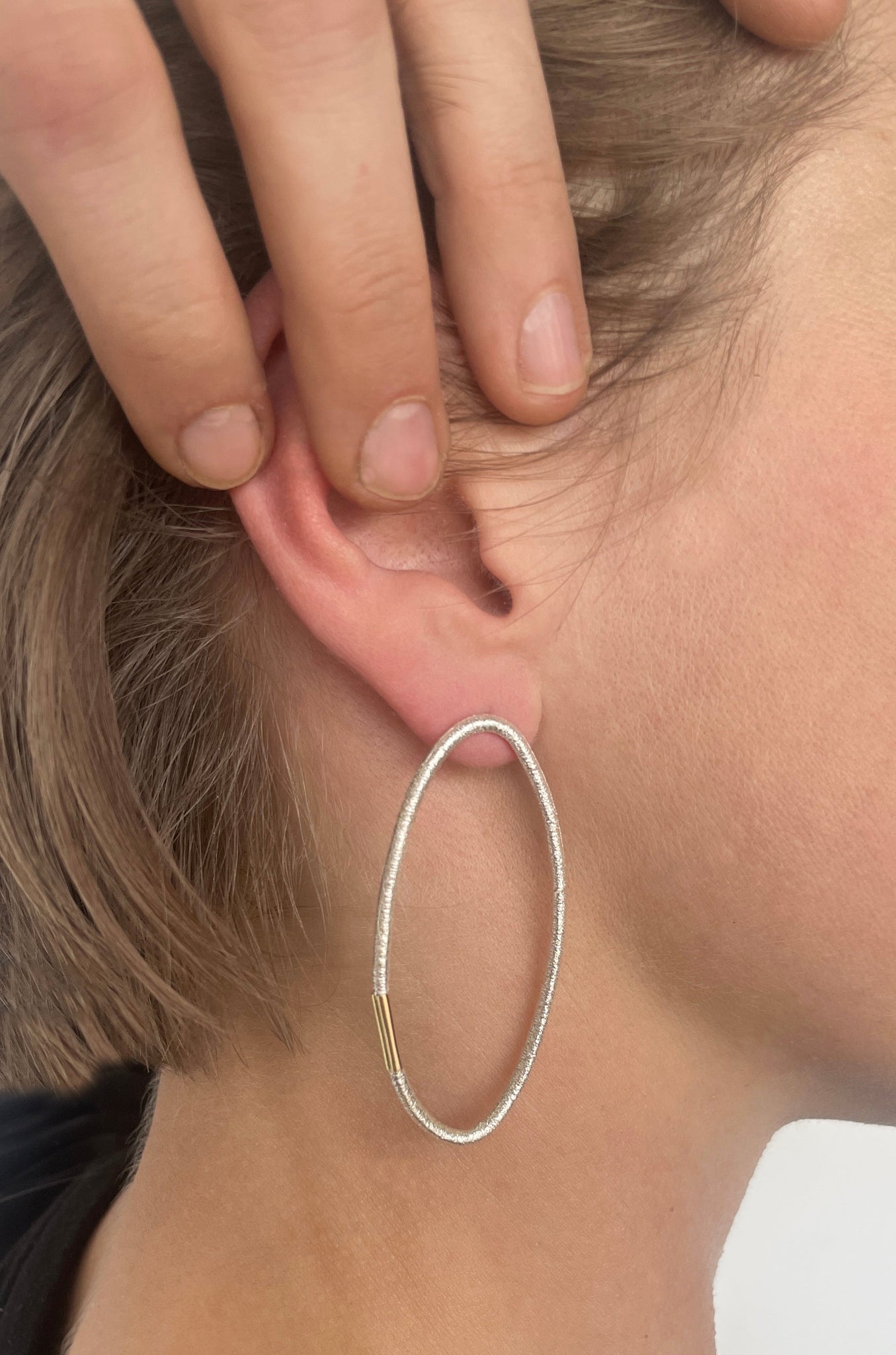 Ohrstecker aus Silber mit ovaler Form und charakteristischer Haargummistruktur, minimalistisch und handgefertigt, getragen am Ohr einer Frau mit blondem Bob.