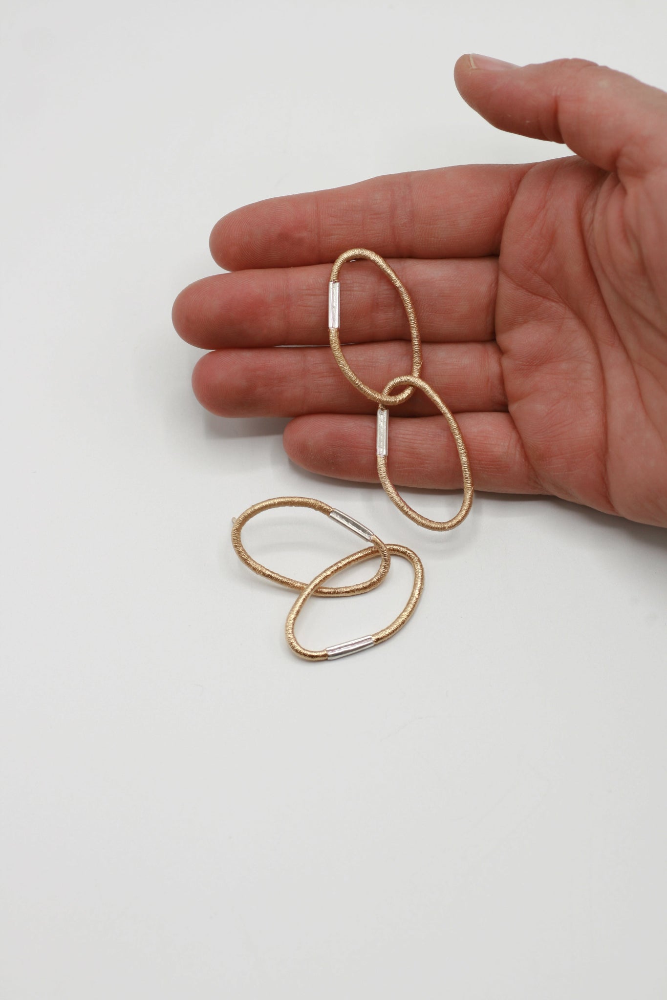 Ein Paar Ohrringe aus je zwei ovalen Gliedern mit charakteristischer Haargummistruktur und Bördelungsdetail an der Schliesse, minimalistisch und handgefertigt. Vergoldetes Silber mit silberner Schliesse, arrangiert auf hellem Hintergrund, ein Paar gehalten von einer Hand.