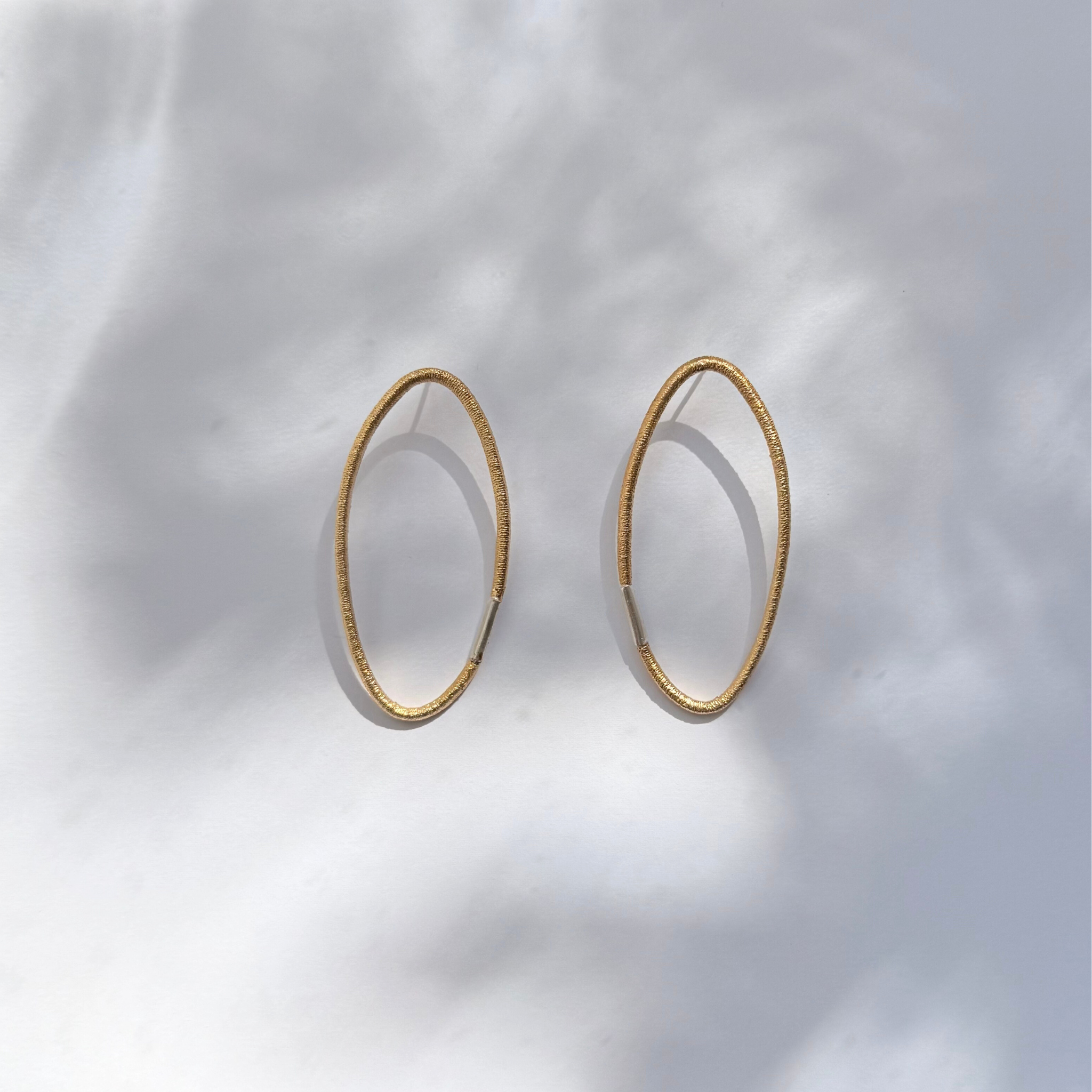Vergoldete Ohrstecker aus Silber mit ovaler Form und charakteristischer Haargummistruktur, minimalistisch und handgefertigt, Bördelung an der Schliesse in Silber, paarweise arrangiert auf hellem Hintergrund.
