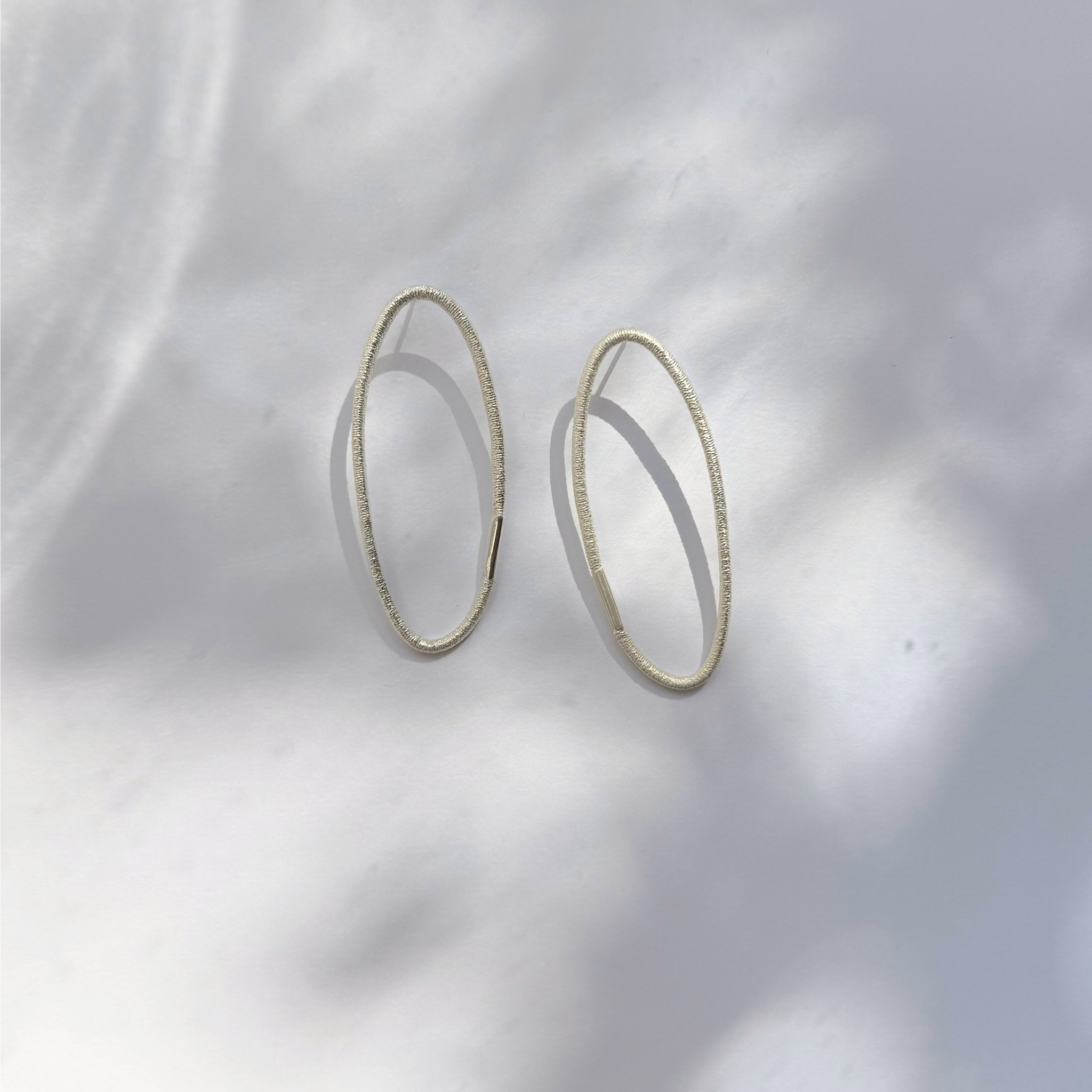 Ohrstecker aus Silber mit ovaler Form und charakteristischer Haargummistruktur, minimalistisch und handgefertigt, paarweise arrangiert auf hellem Hintergrund.