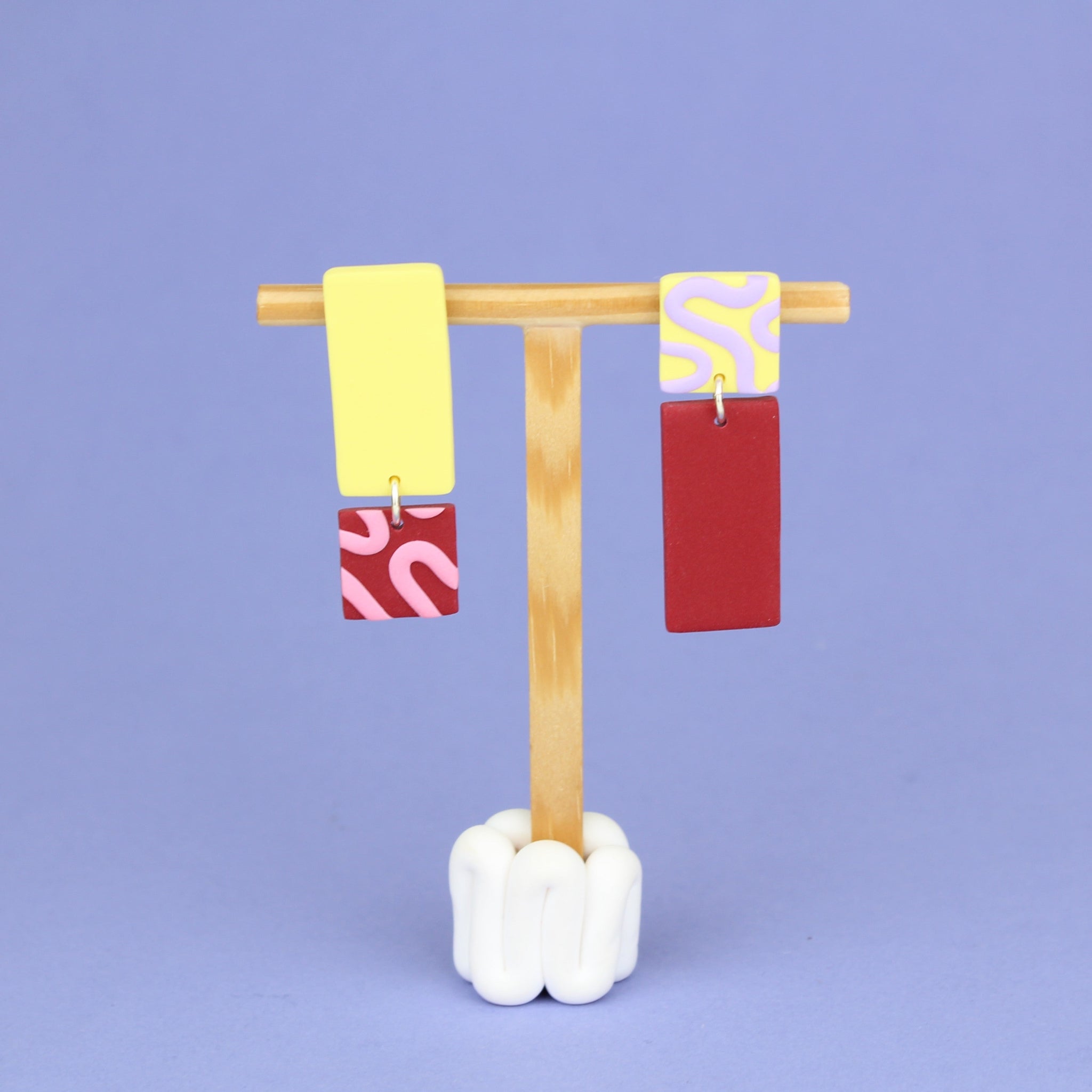Asymmetrische Ohrringe aus Polymerton mit rechteckigen Elementen und grafischen Mustern in Gelb, Rot, Rosa und Flieder, präsentiert auf einem hölzernen Schmuckständer.