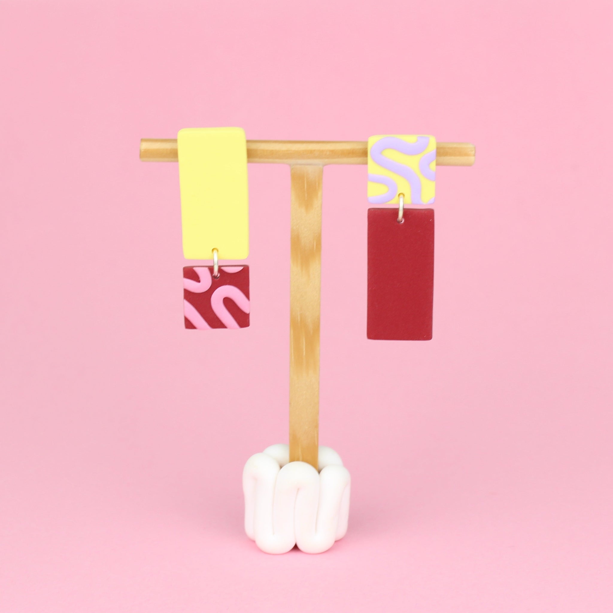 Asymmetrische Ohrringe aus Polymerton mit rechteckigen Elementen und grafischen Mustern in Gelb, Rot, Rosa und Flieder, präsentiert auf einem hölzernen Schmuckständer.