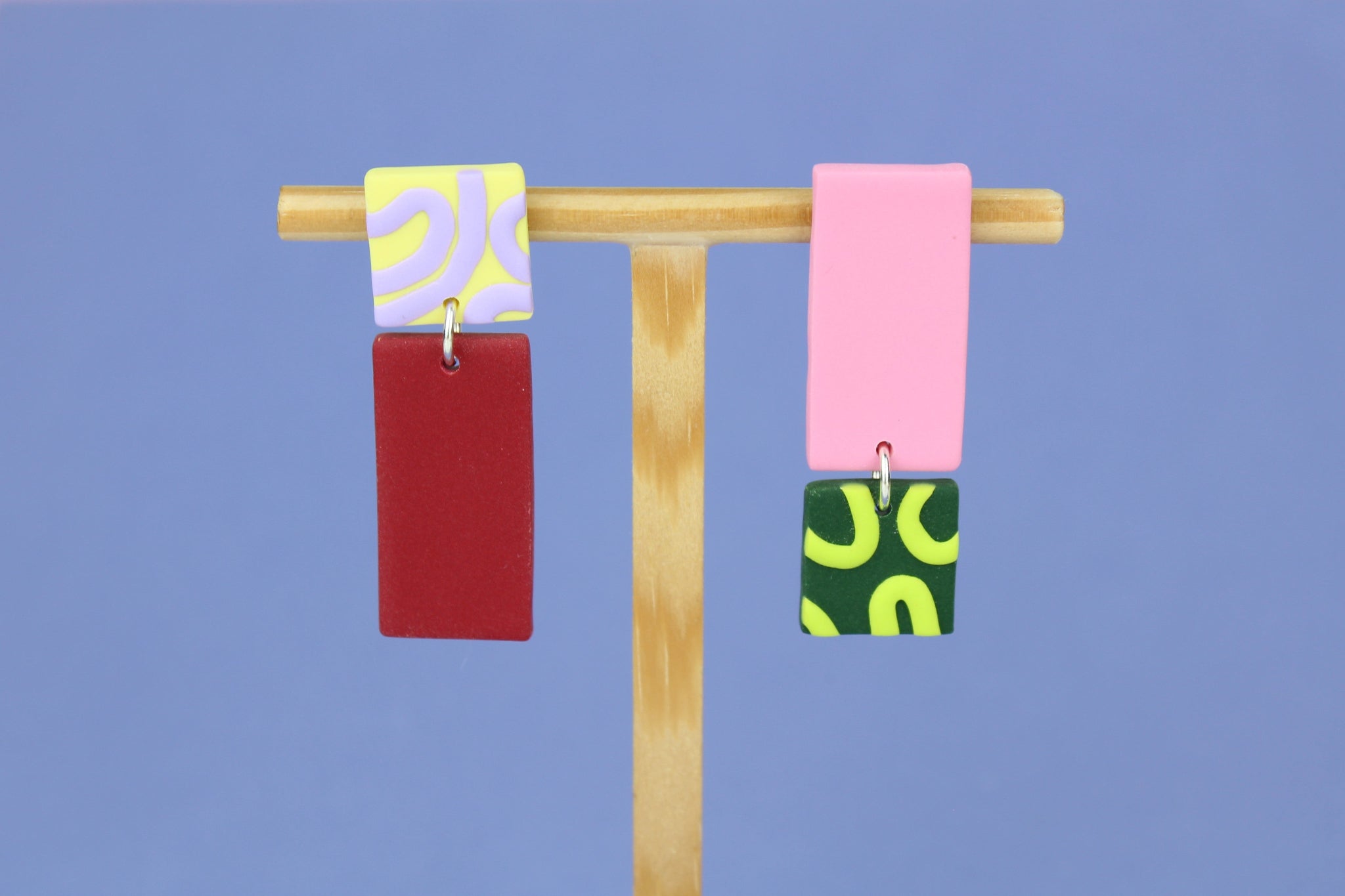 Asymmetrische Ohrringe aus Polymerton mit rechteckigen Elementen und grafischen Mustern in Rot, Rosa, Buttergelb, Grün und Flieder, präsentiert auf einem hölzernen Schmuckständer.
