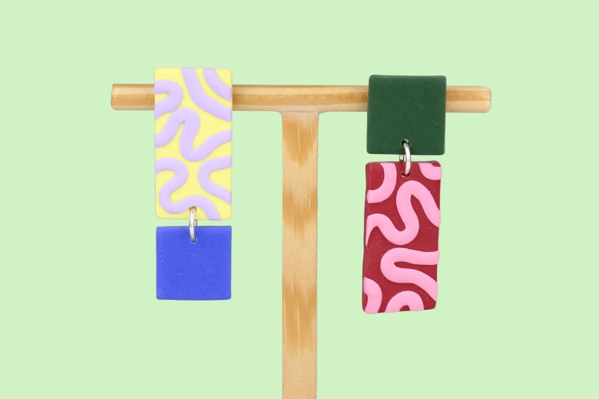 Asymmetrische Ohrringe aus Polymerton mit rechteckigen Elementen und grafischen Mustern in Gelb, Rot, Flieder, Blau und Grün, präsentiert auf einem hölzernen Schmuckständer.