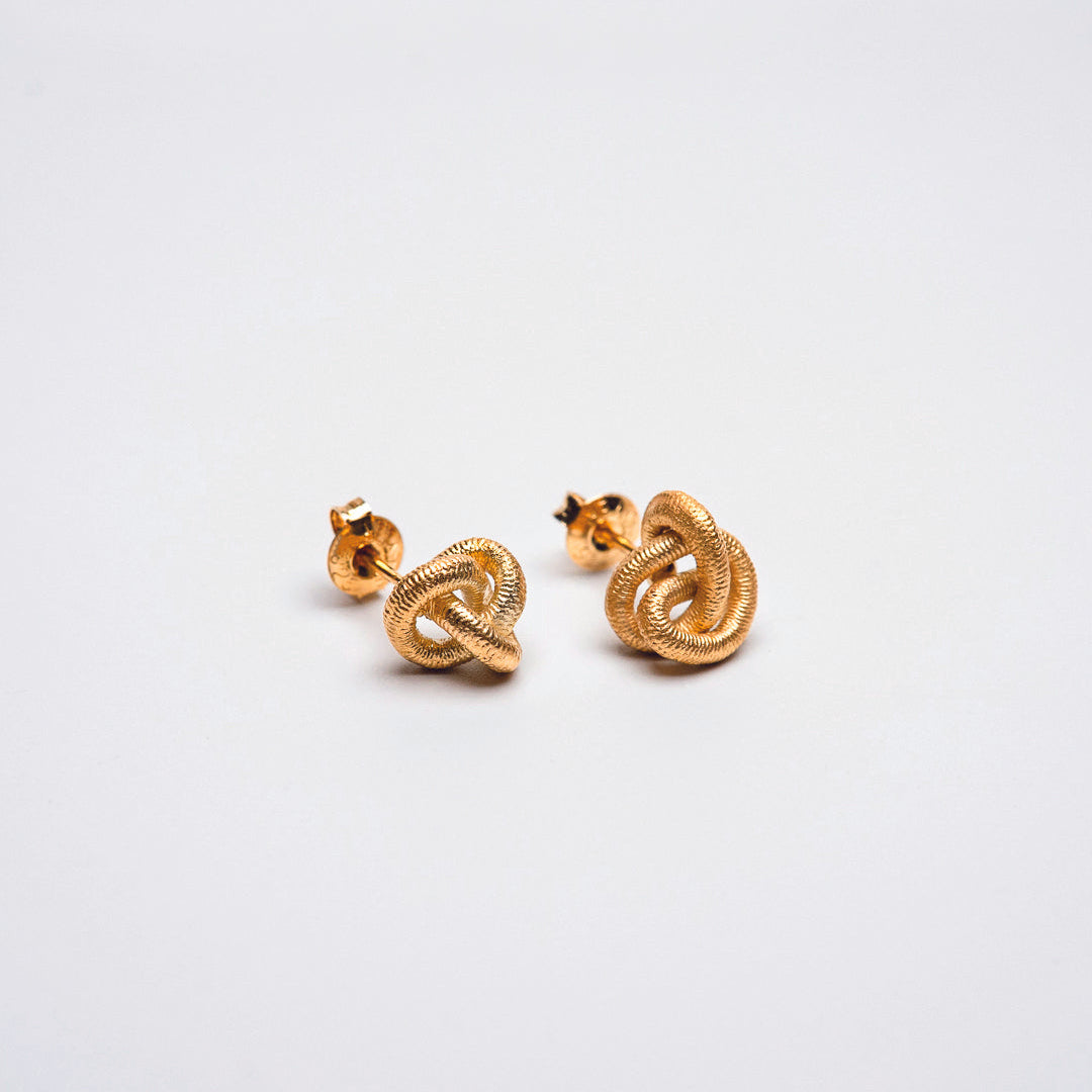 Paar goldfarbene Ohrstecker in knotiger, organischer Form, auf hellem Hintergrund fotografiert. Die strukturierte Oberfläche verleiht den kleinen Skulptur-Ohrringen eine textile Anmutung.