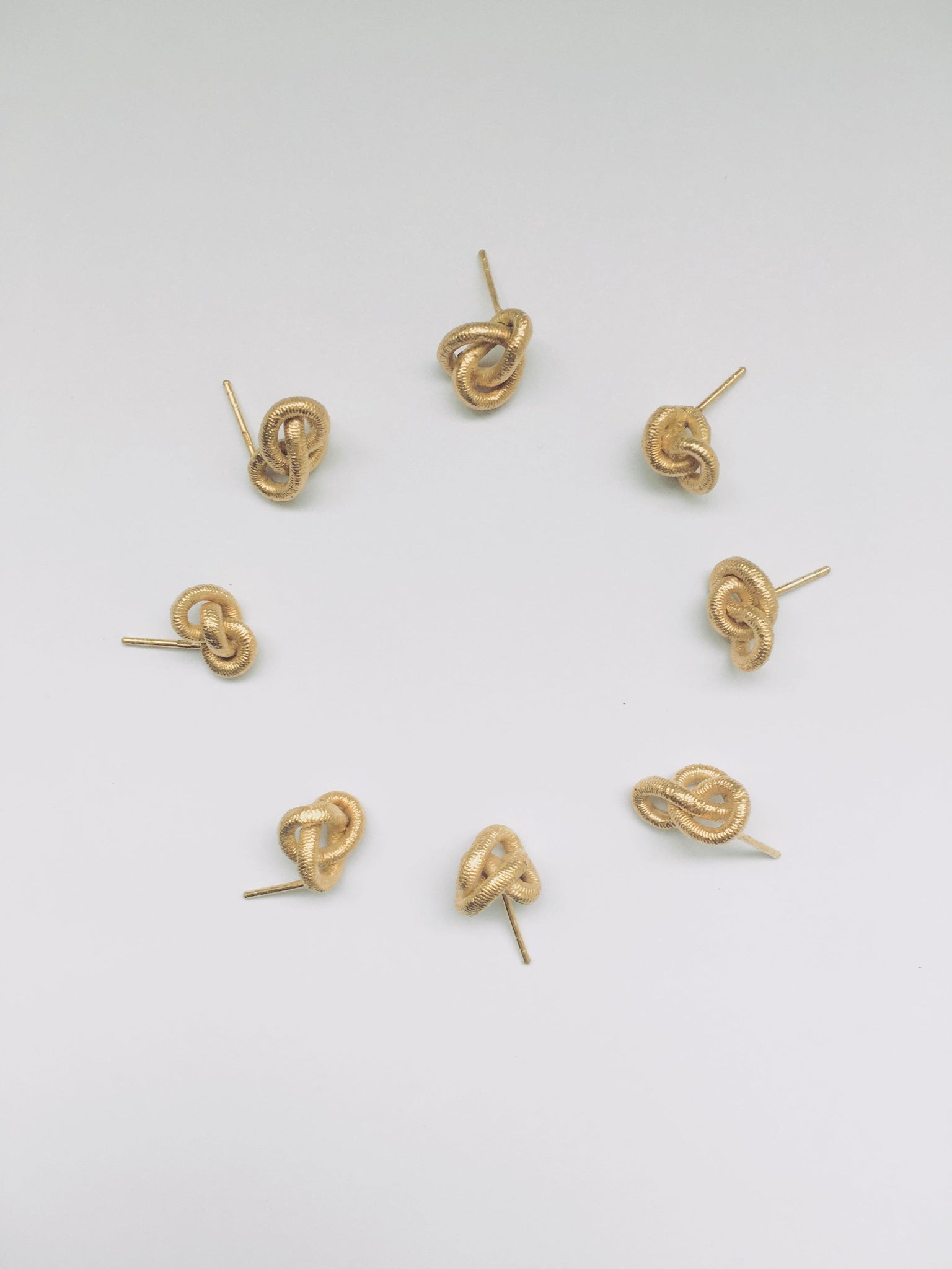 Acht kleine goldene Ohrstecker in unterschiedlichen Knotenform liegen in einem Kreis auf einem weissen Studiohintergrund.