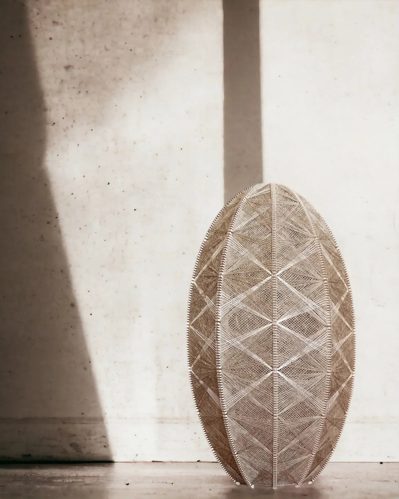 ein eiförmiger gewebter Lampenschirm aus Birkenholz und Leinengarn steht vor einer schattigen Wand