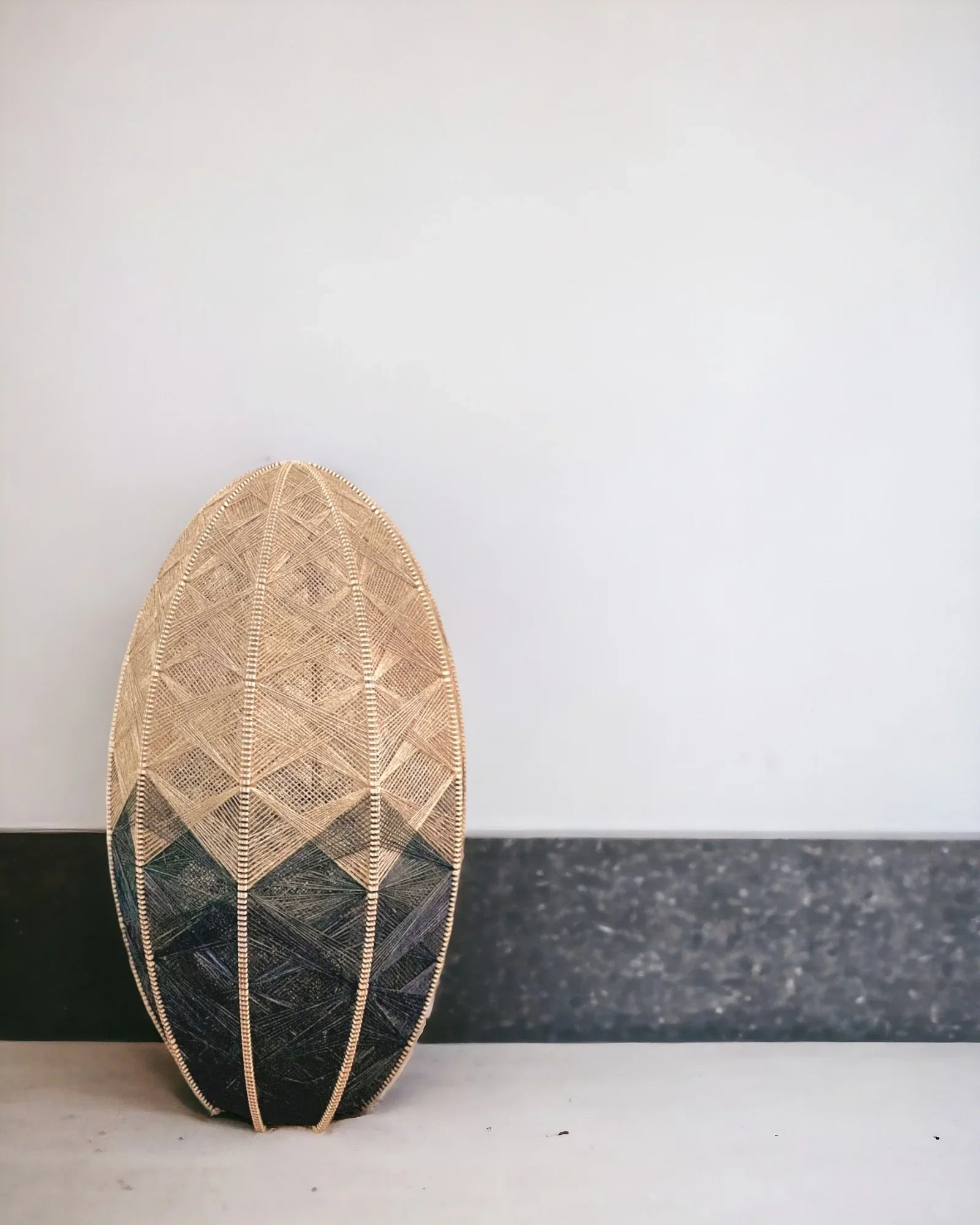 eine zylindrisch Stehlampe aus einem Birkenholzrahmen umwebt mit Leinengarn steht vor einer weißen Wand mit schwarzem Sockel