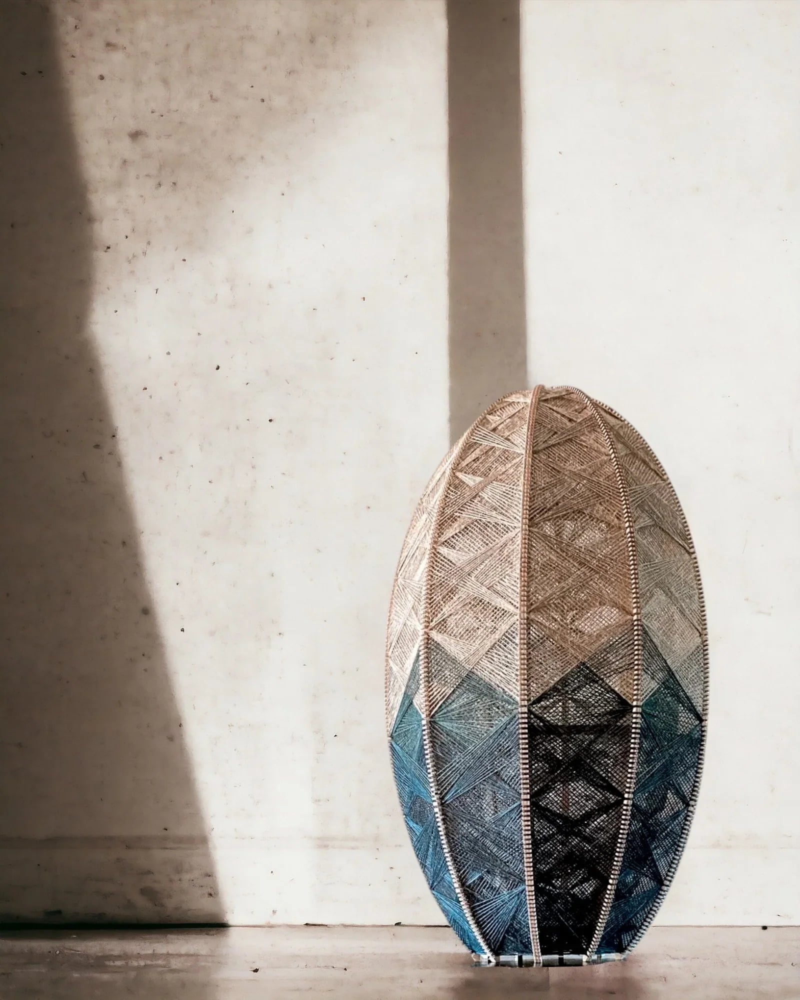 eine handgewebte Lampe aus Birkenholz und Leinengarn in Beige und Anthrazit steht vor einer mediterranen Wand