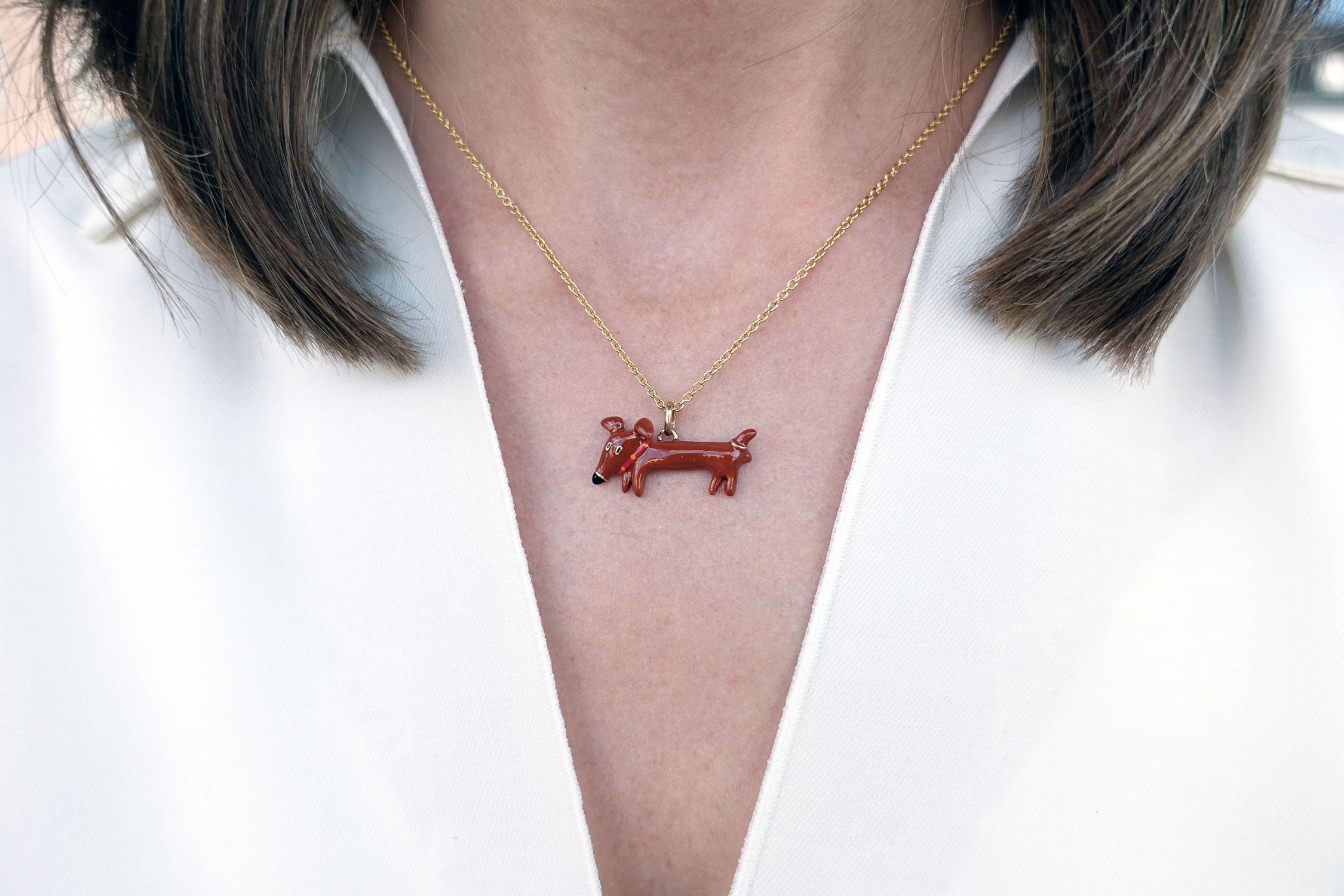 Eine schmale goldene Halskette mit einem Anhänger in Form eines braunen Dackels mit rotem Halsband, schwarzer Schnauzen und goldenen Konturen der italienischen Schmuckdesignerin Chiara Bello von chic pig.