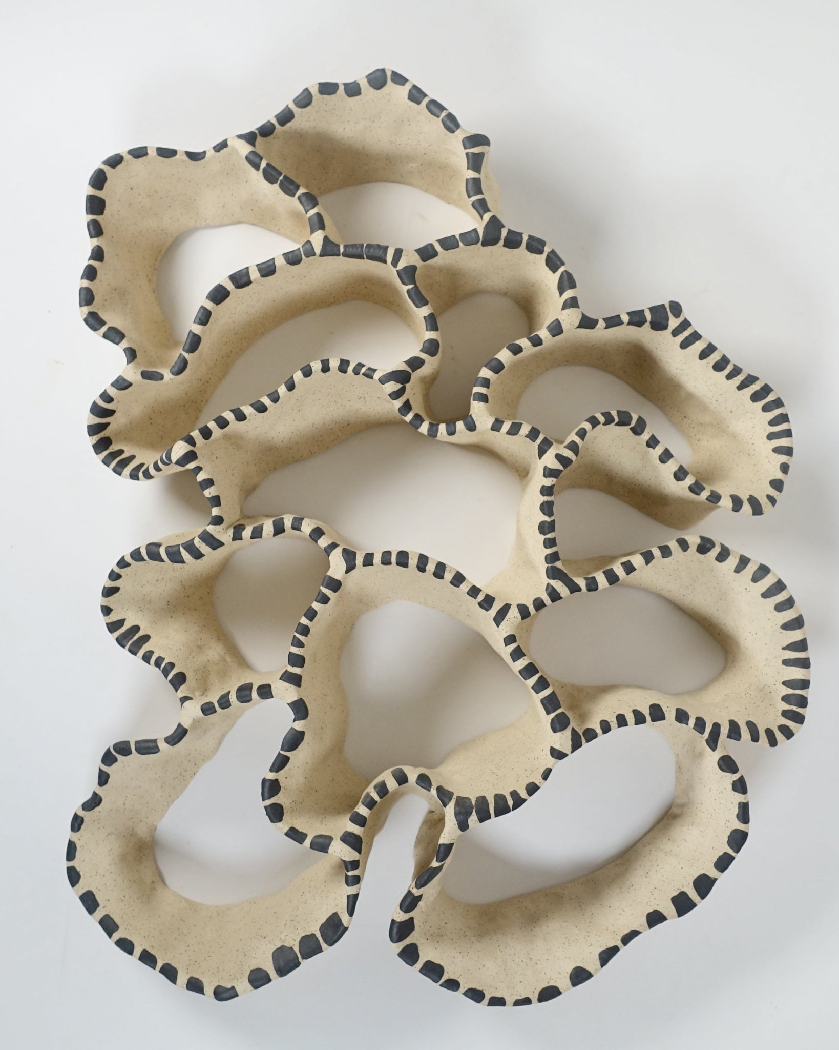 Keramische Wandskulptur mit offenem, durchbrochenem Aufbau, angelehnt an eine Korallenform; sandfarbene Oberfläche mit unregelmäßig geschwungenen Formen, entlang der Kanten mit rhythmisch gesetzten, dunkelgrauen Pinselstrichen akzentuiert.