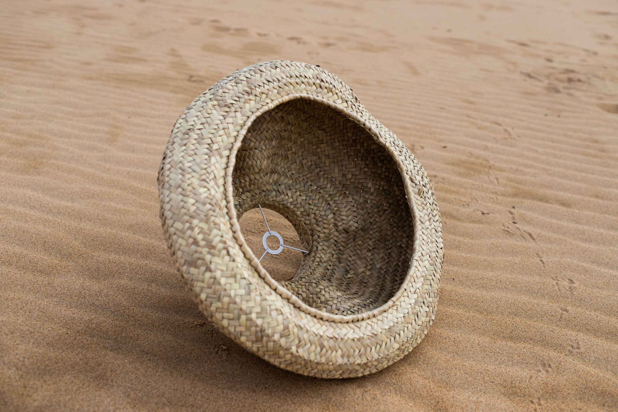 Unteransicht des handgewebten Lampenschirms DUNA I von Ysaline Ophoff aus empreita de palma, organisch-dünenförmig geflochten, auf hellem Sand liegend, man kann durch die untere Öffnung die metallene Halterung für die Fassung sehen.