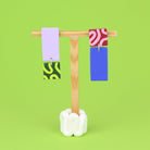 Asymmetrische Ohrringe aus Polymerton mit rechteckigen Elementen und grafischen Mustern in Rosa und Grün, präsentiert auf einem hölzernen Schmuckständer vor hellgrünem Hintergrund.