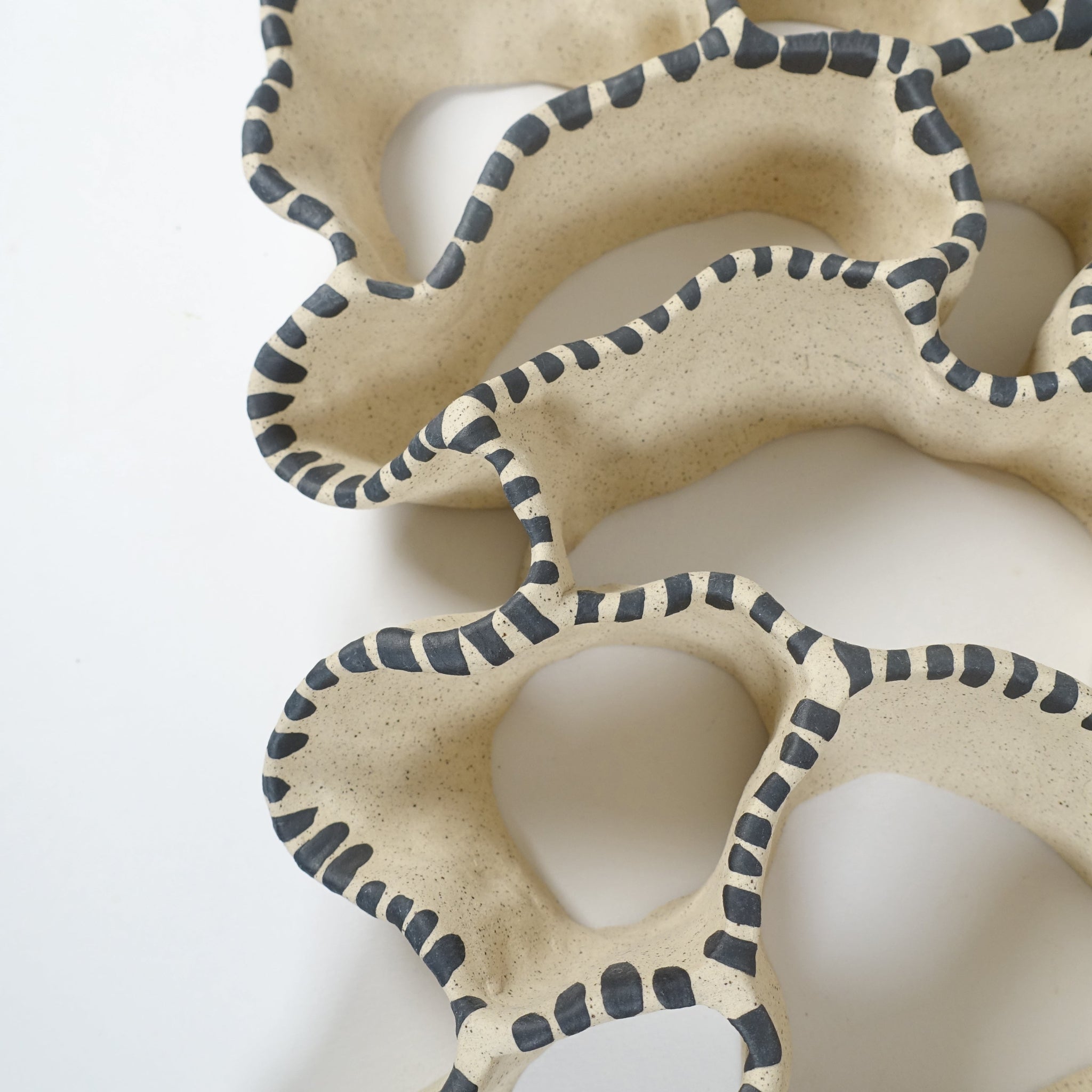 Detailansicht einer keramischen Wandskulptur mit offenem, durchbrochenem Aufbau, angelehnt an eine Korallenform; sandfarbene Oberfläche mit unregelmäßig geschwungenen Formen, entlang der Kanten mit rhythmisch gesetzten, schwarzen Pinselstrichen akzentuiert.