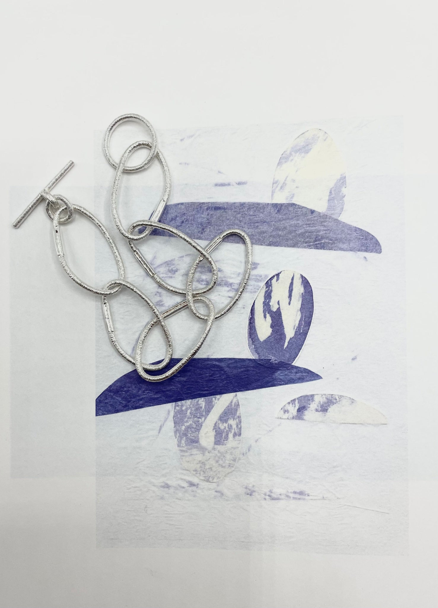 Versilbertes Armband aus Gliedern, die von Haargummis abgegossen sind, mit T-Verschluss, arrangiert auf einem grafischen Untergrund in Blau- und Weißtönen.