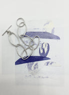 Versilbertes Armband aus Gliedern, die von Haargummis abgegossen sind, mit T-Verschluss, arrangiert auf einem grafischen Untergrund in Blau- und Weißtönen.