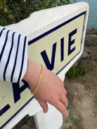 Vermeil-Armreif aus vergoldetem Silber mit Haargummistruktur, minimalistisch aber mit Bördelungsdetail an der Schliesse, getragen am Handgelenk vor maritimem Wegweiser im Außenbereich.