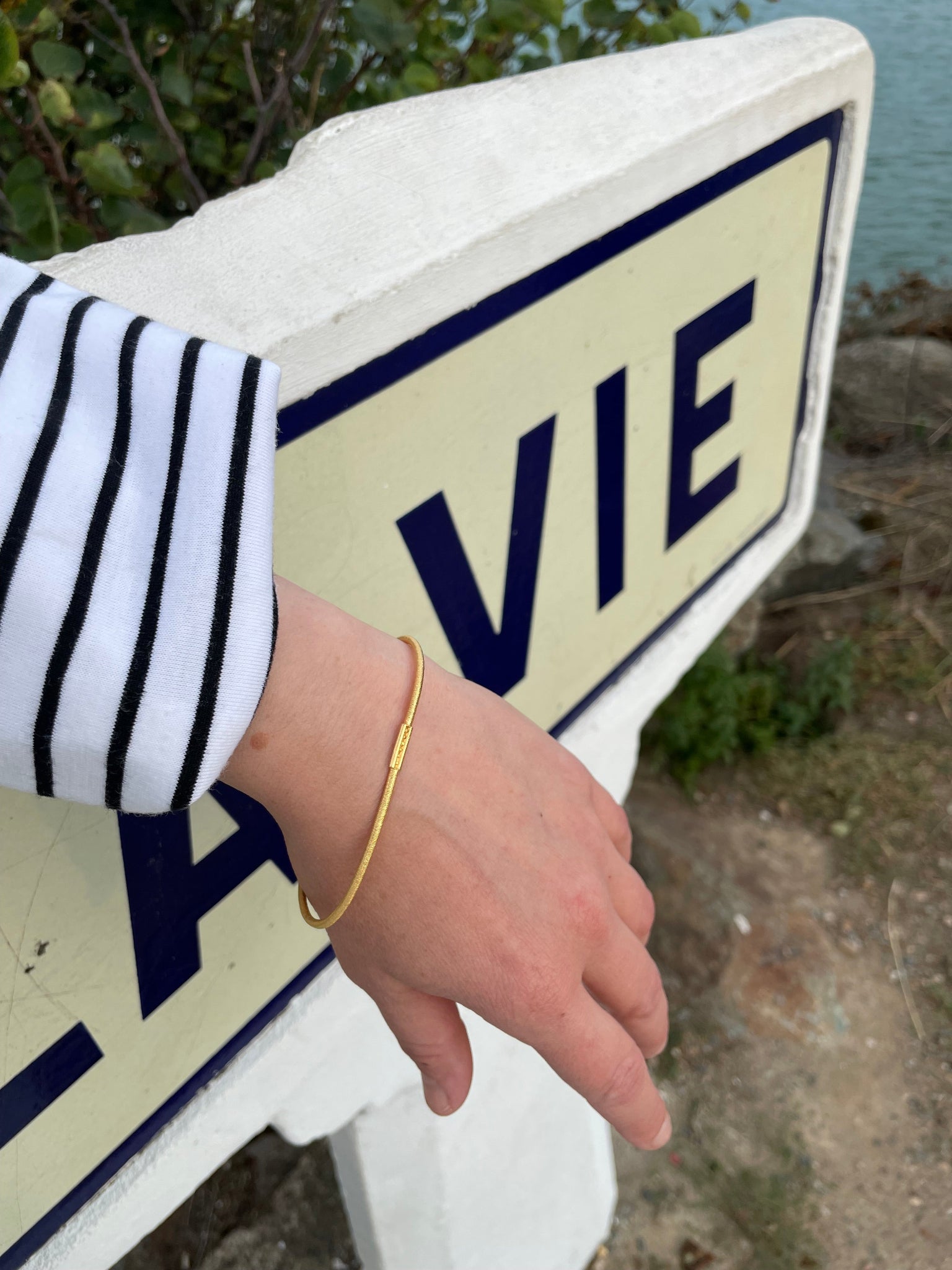 Vermeil-Armreif aus vergoldetem Silber mit Haargummistruktur, minimalistisch aber mit Bördelungsdetail an der Schliesse, getragen am Handgelenk vor maritimem Wegweiser im Außenbereich.