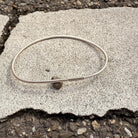 Silberner Armreif mit Haargummistruktur und Bördelung an der Schliesse, minimalistisch und handgefertigt, liegend auf hellem Steinuntergrund im Außenbereich.