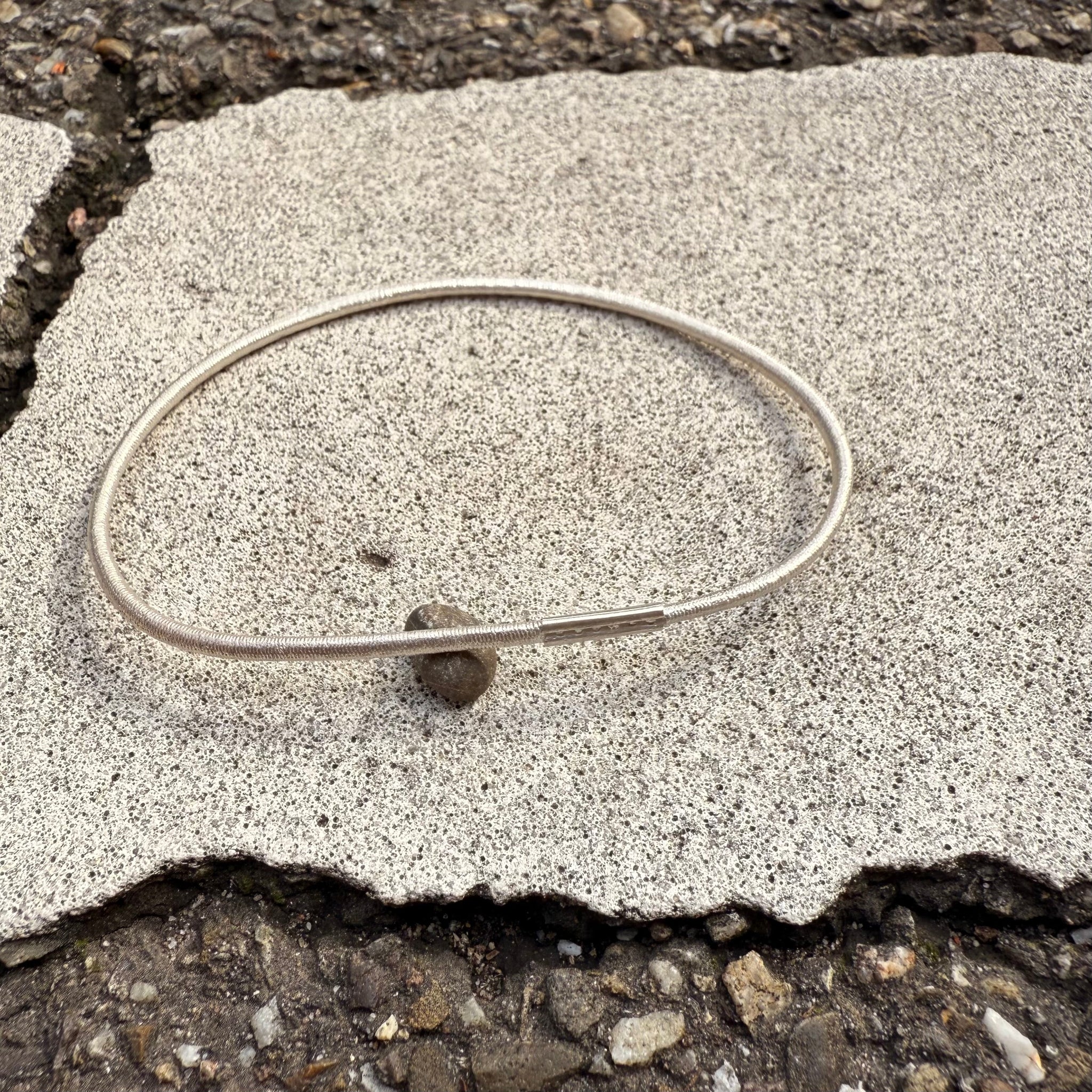 Silberner Armreif mit Haargummistruktur und Bördelung an der Schliesse, minimalistisch und handgefertigt, liegend auf hellem Steinuntergrund im Außenbereich.