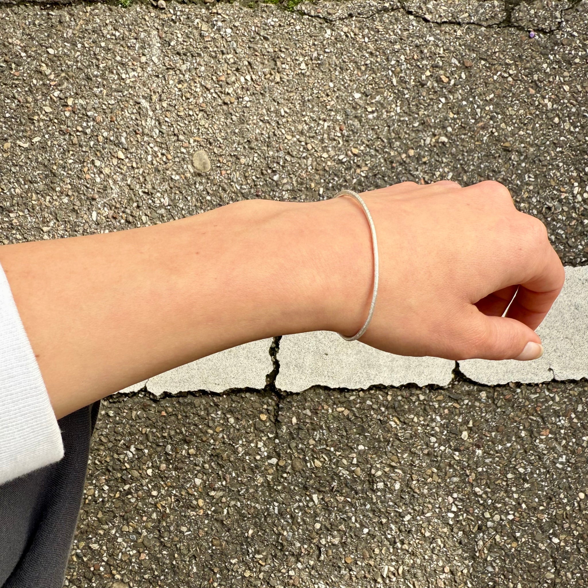 Silberner Armreif mit Haargummistruktur und Bördelung an der Schliesse, minimalistisch und handgefertigt, getragen an einem Handgelenk mit weissem Pulli.