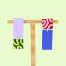 Asymmetrische Ohrringe aus Polymerton mit rechteckigen Elementen und grafischen Mustern in Flieder und Blau, präsentiert auf einem hölzernen Schmuckständer.